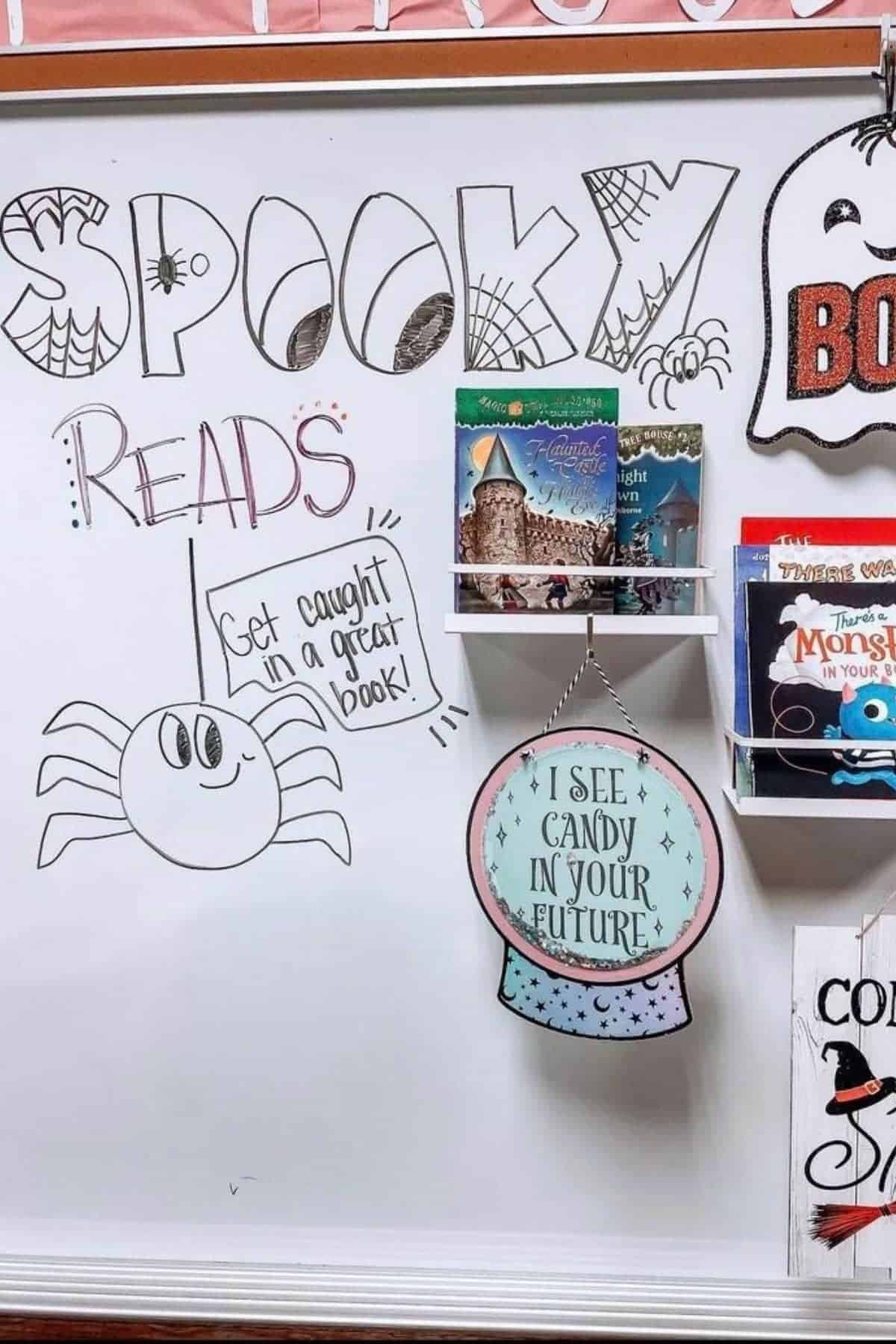 Whiteboard decorated with spider and ghost drawings, displaying books on a small shelf with the text “Spooky Reads” and “I see candy in your future” on a hanging sign.