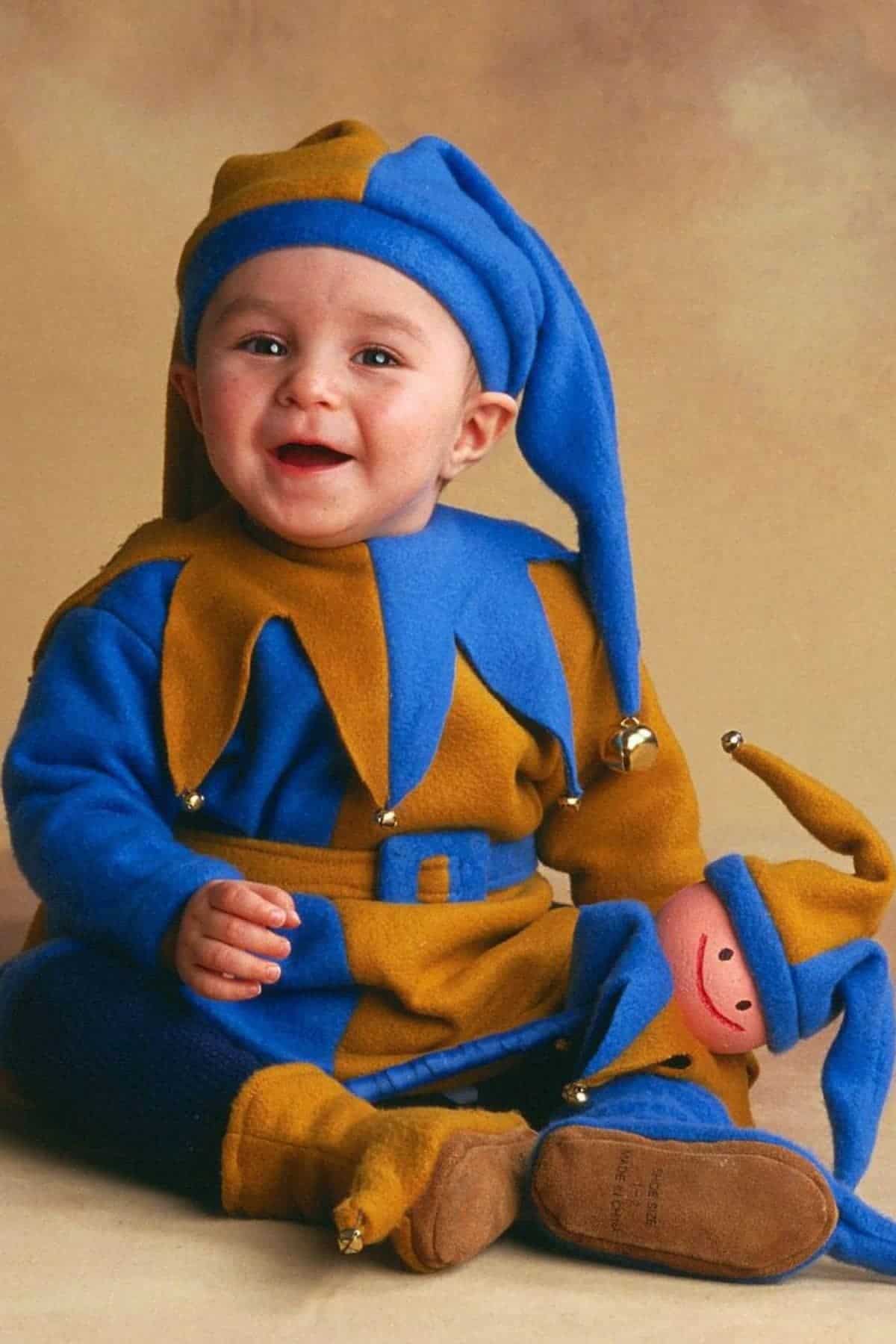 A baby dressed in a blue and gold jester costume sits and smiles, holding a matching jester doll.