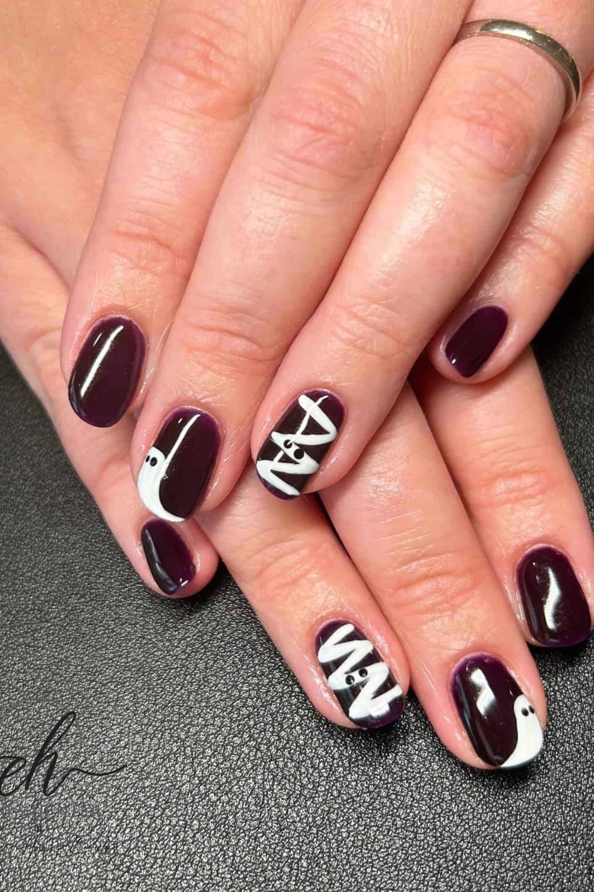 Hands with short, dark purple nails featuring white Halloween-themed designs including a mummy, eyeballs, and bandages.