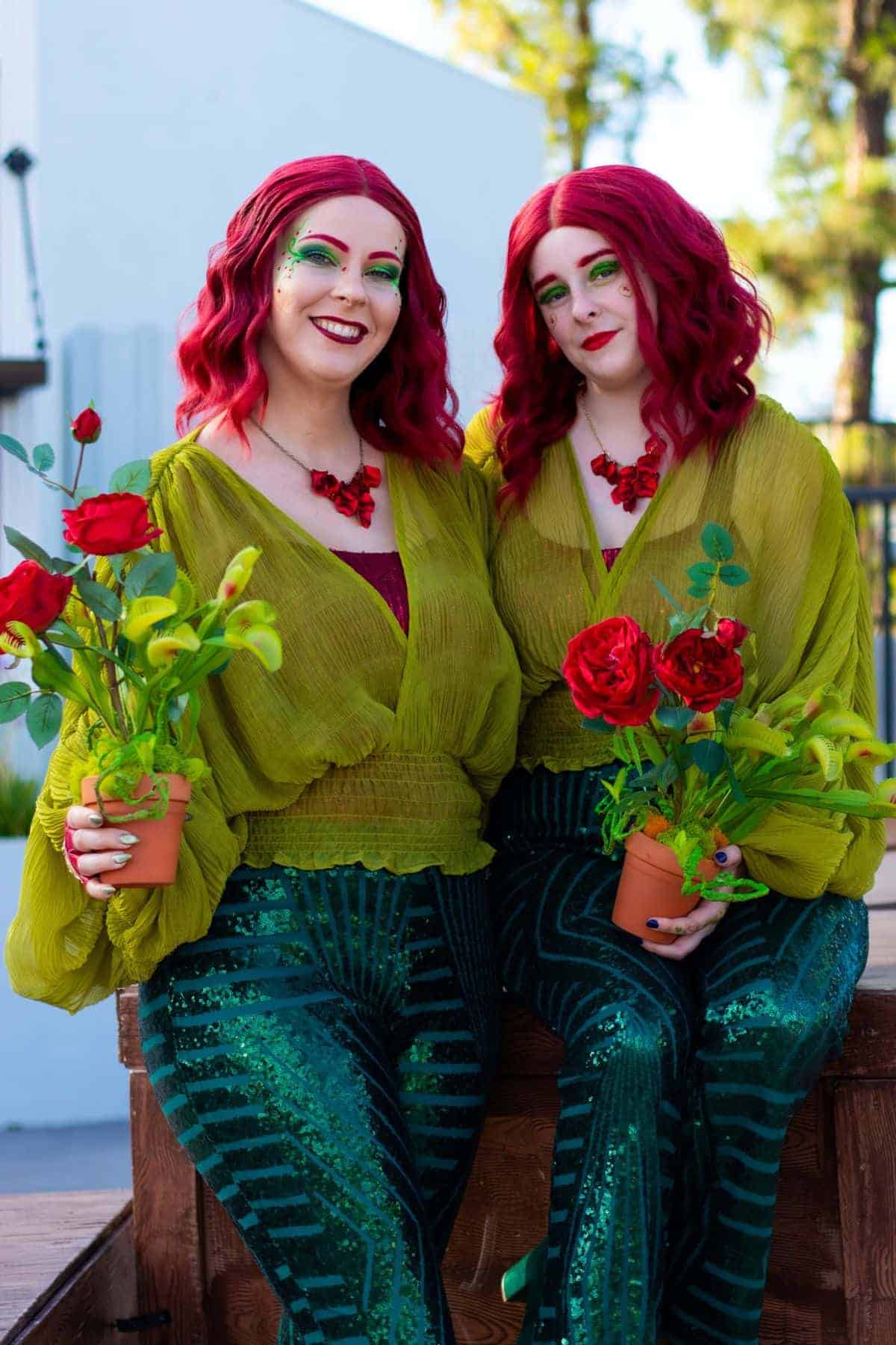 Two people with red hair, green eye makeup, and matching green outfits hold potted red roses and smile outdoors.