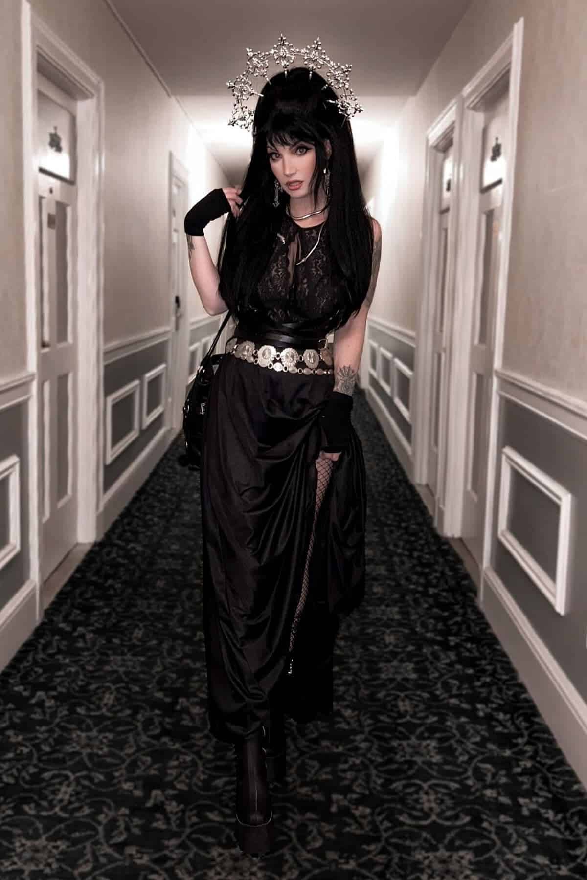 Person in a black gothic outfit with a silver crown stands in a carpeted hallway with white doors and walls, looking at the camera.