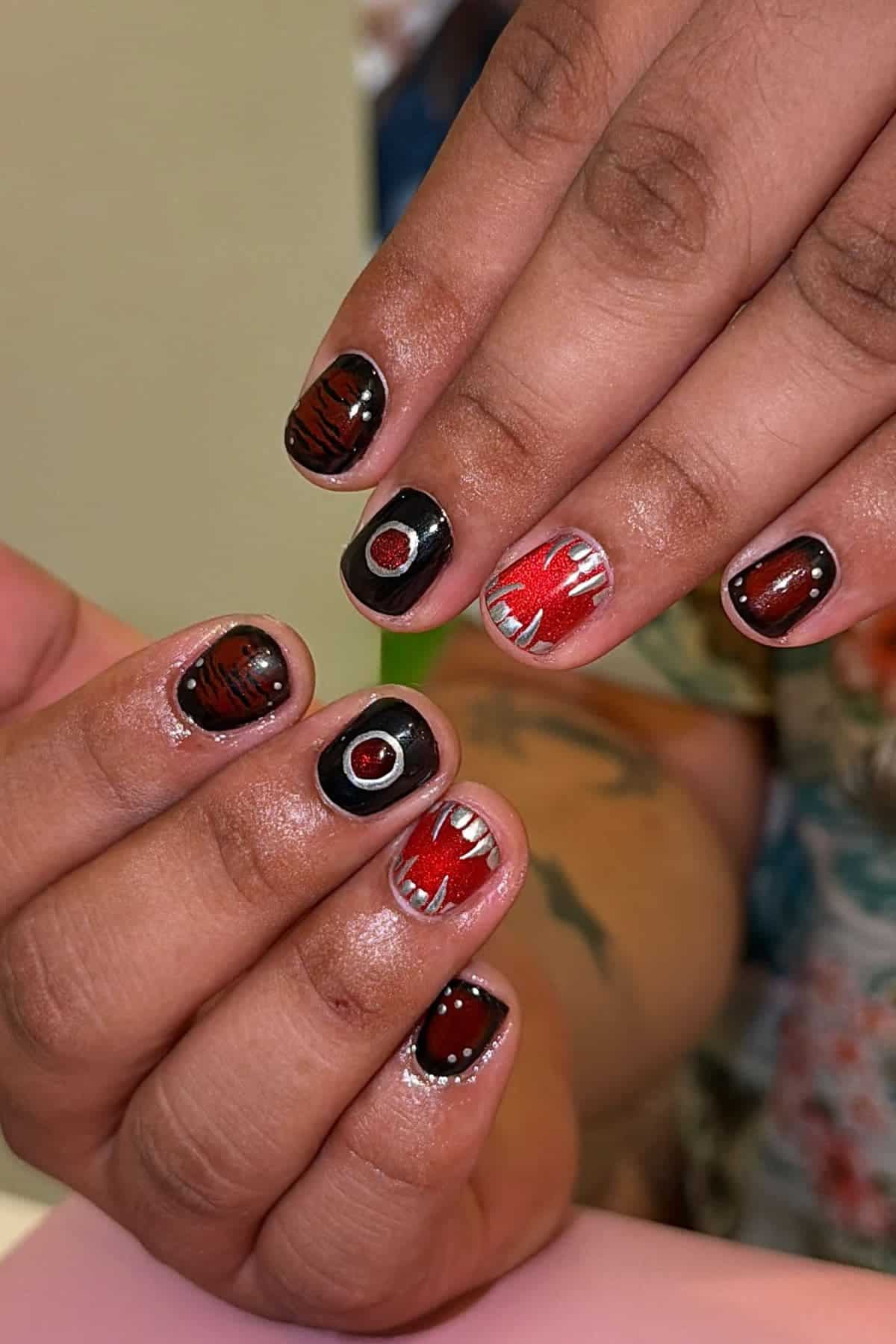 Close-up of hands with short nails painted in black and red designs, including eyes, teeth, and abstract patterns. The person has a tattoo visible on their arm.