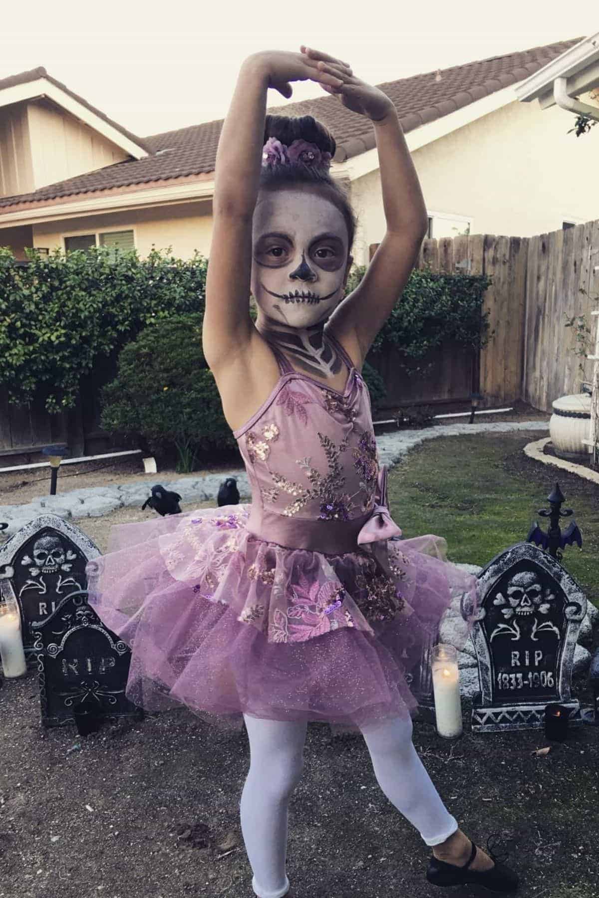 A young girl in a purple ballet costume and skeleton face paint poses with arms raised in a yard decorated with Halloween tombstones and candles.