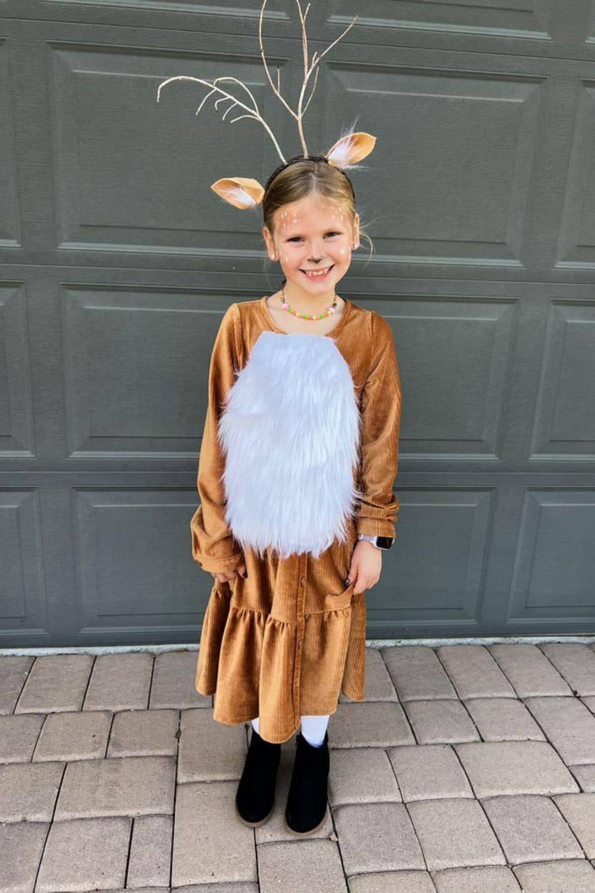 Young child in a deer costume, featuring antlers, ears, a brown dress, white faux fur, and black boots, standing on a paved surface in front of a closed garage door.