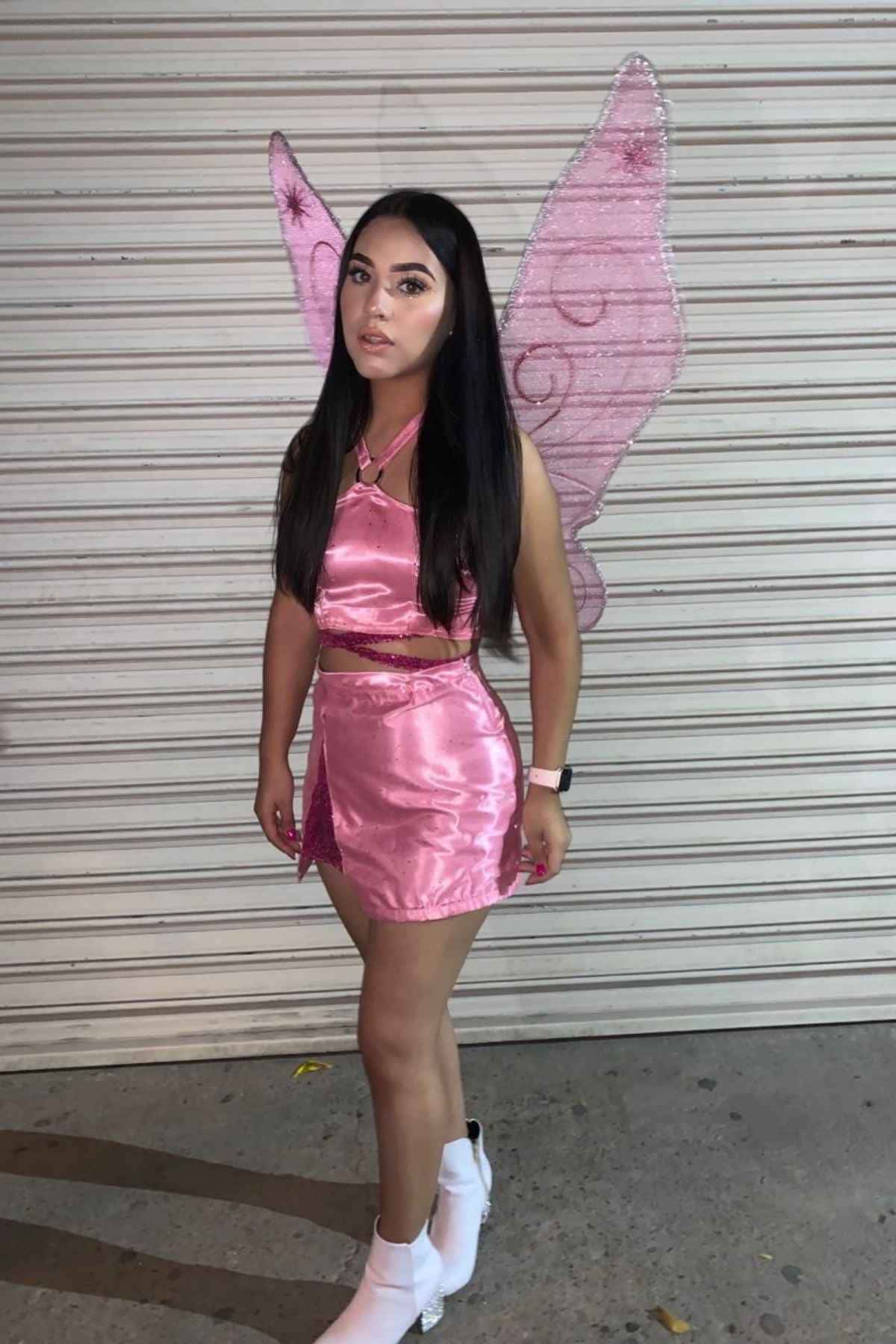 Person wearing a shiny pink outfit with fairy wings and white ankle boots, standing in front of a closed metal shutter at night.