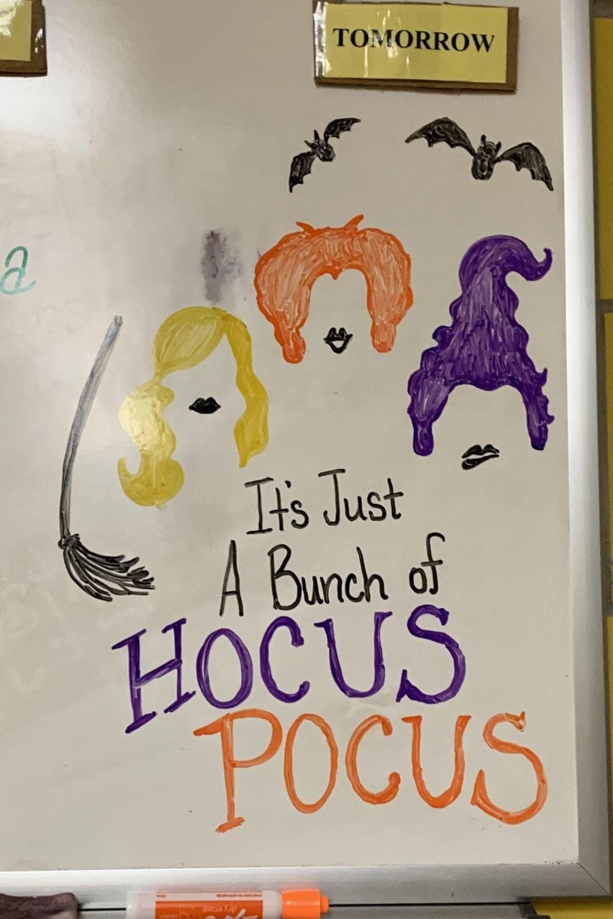 Drawing of three stylized witches’ hairstyles, two bats, and a broom above the phrase “It’s Just A Bunch of Hocus Pocus” on a whiteboard.