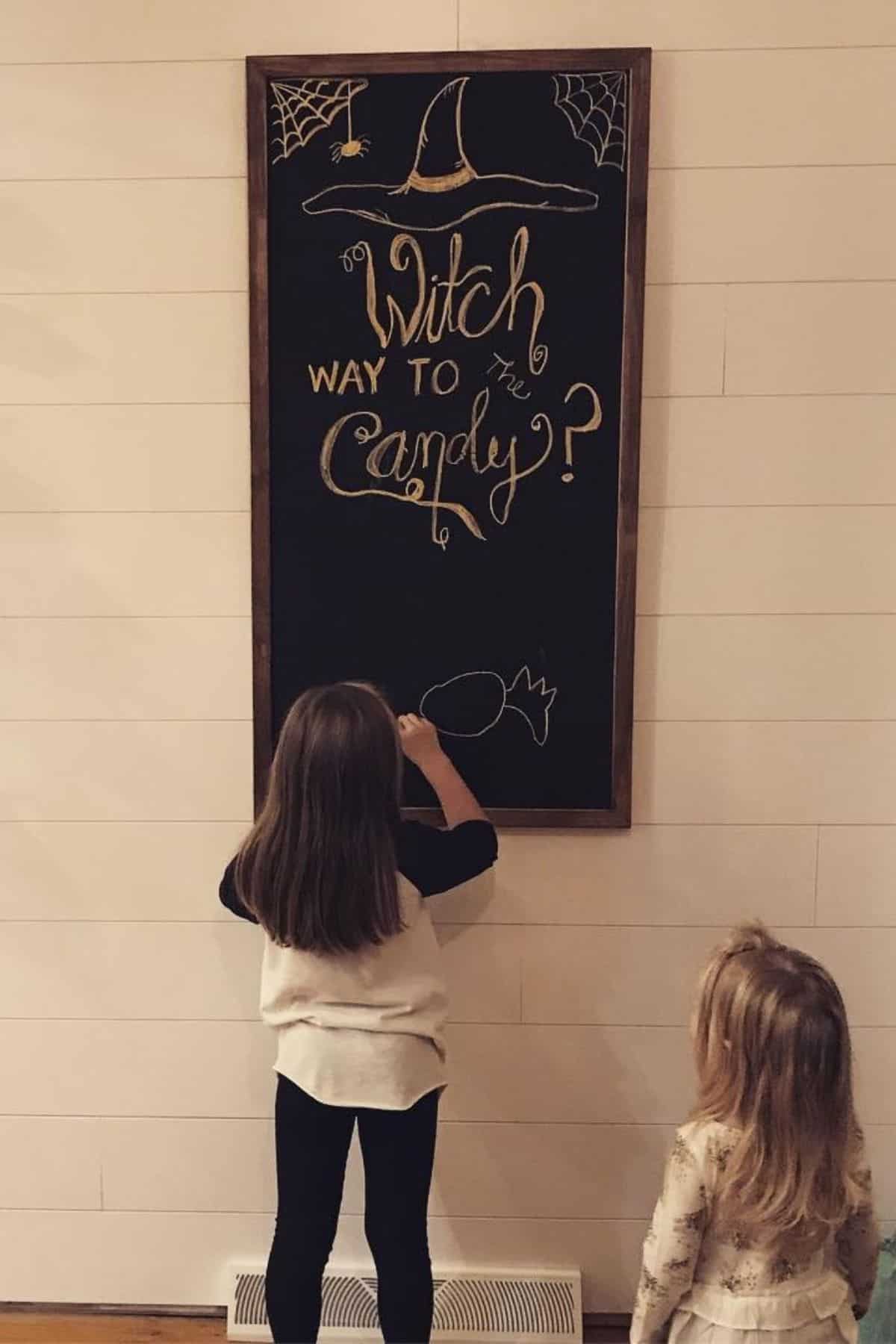 Two young girls stand in front of a chalkboard with a Halloween drawing and the words “Witch way to the candy?” written on it.