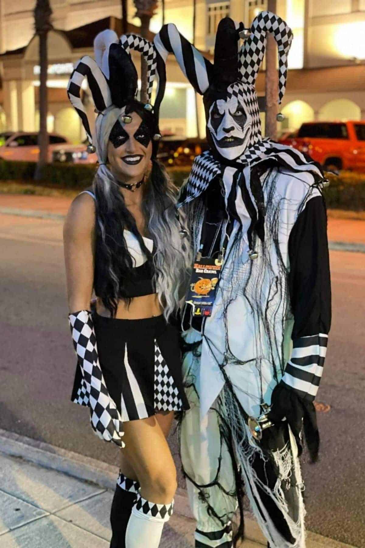 Two people in black-and-white harlequin jester costumes stand together on a city sidewalk at night, with cars and a lit building in the background.