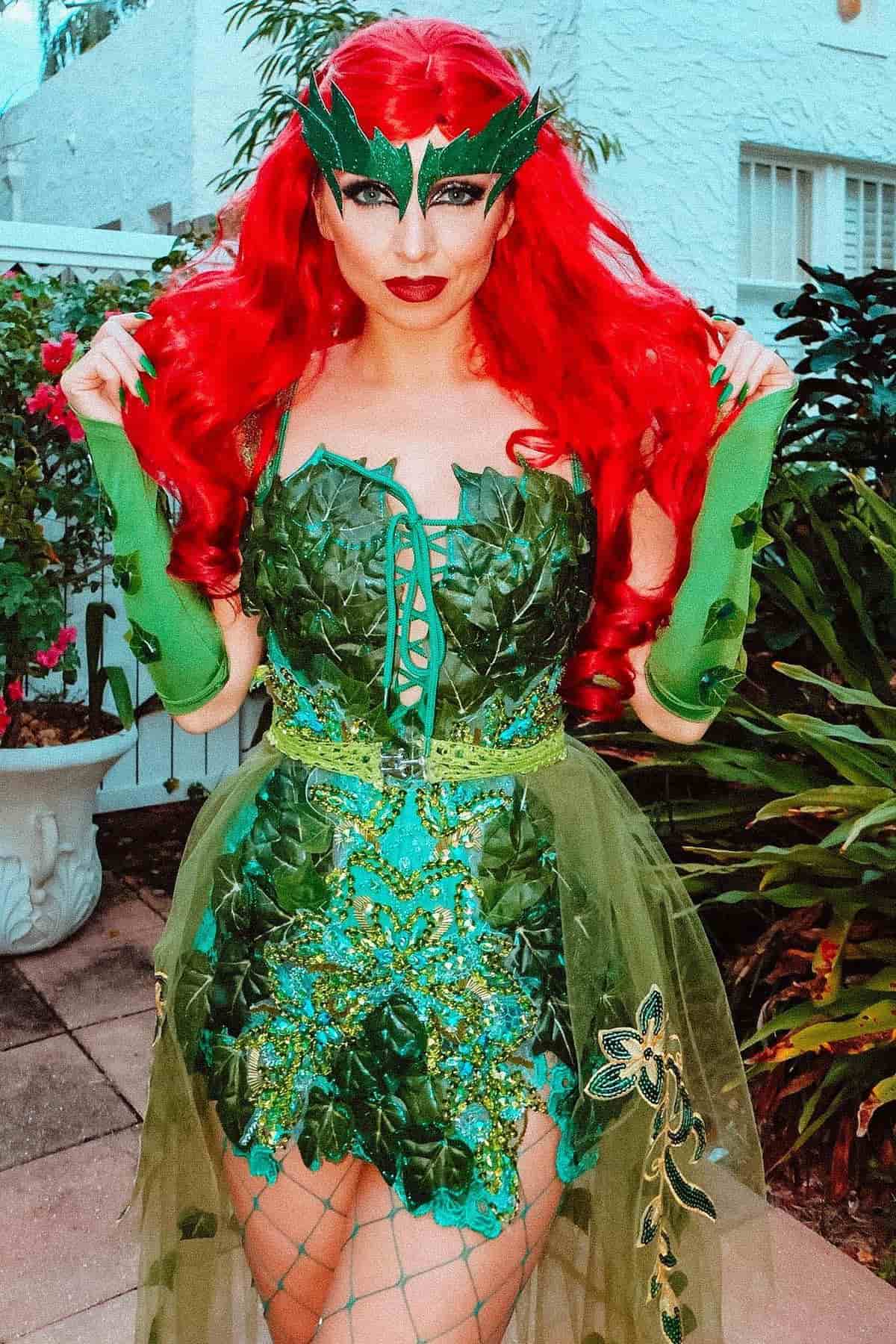 Person in a green leafy costume with red hair, leaf mask, and green gloves stands outdoors near plants and a white building.