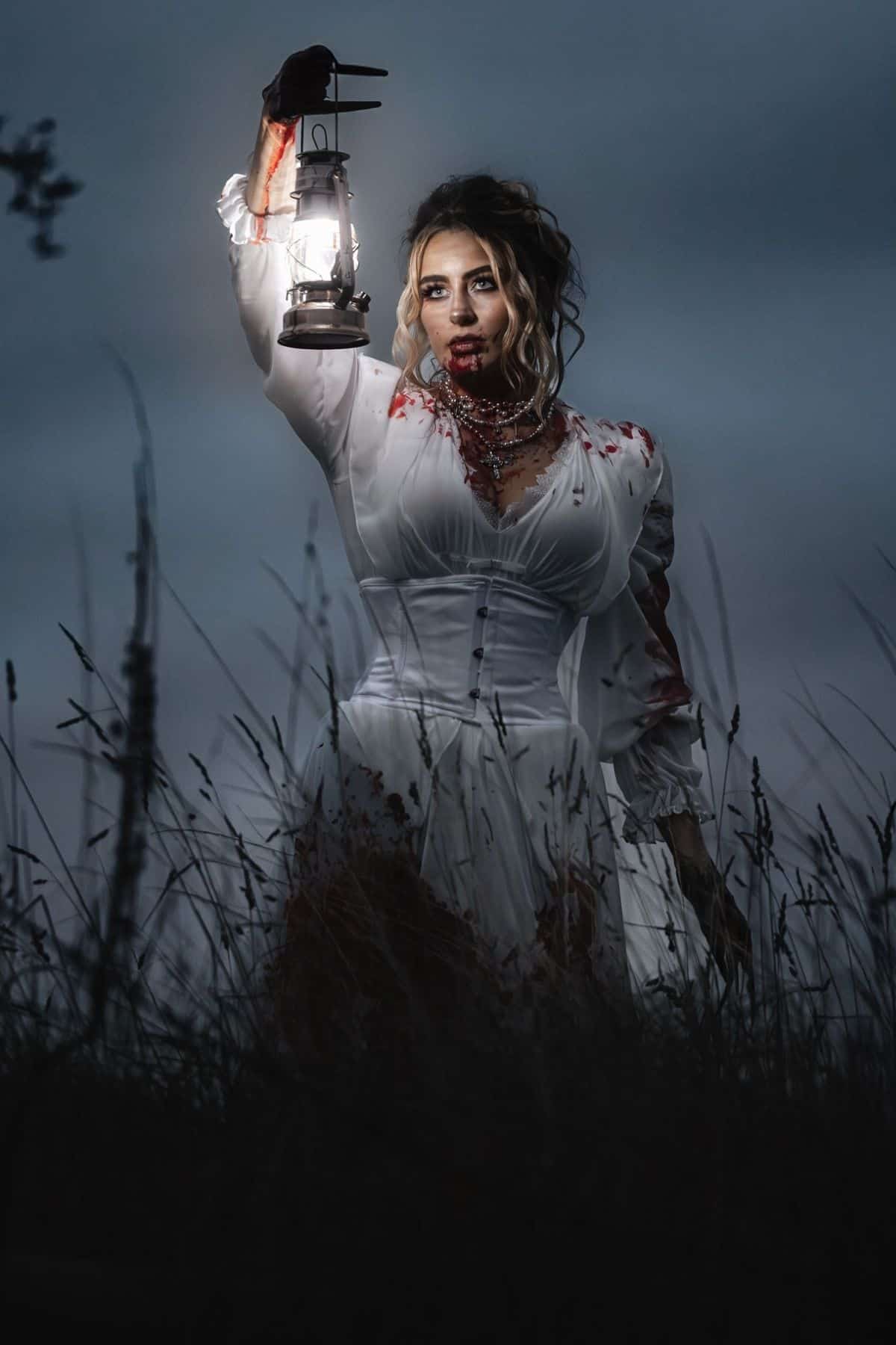 A woman in a blood-stained white dress holds a lit lantern in one hand, standing in tall grass at dusk with a serious expression.