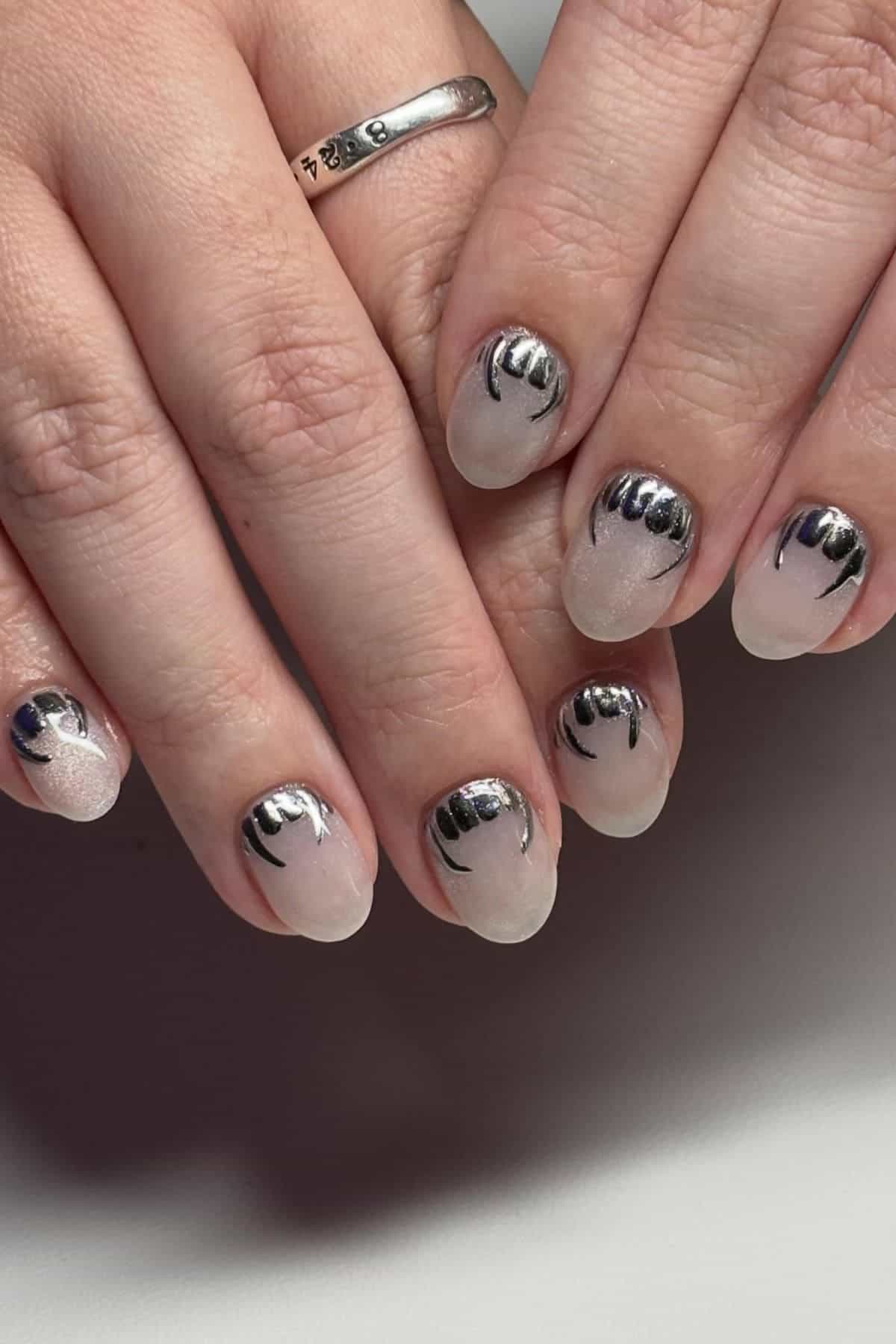 Two hands with short, rounded nails featuring a translucent base and black metallic abstract designs near the cuticles.