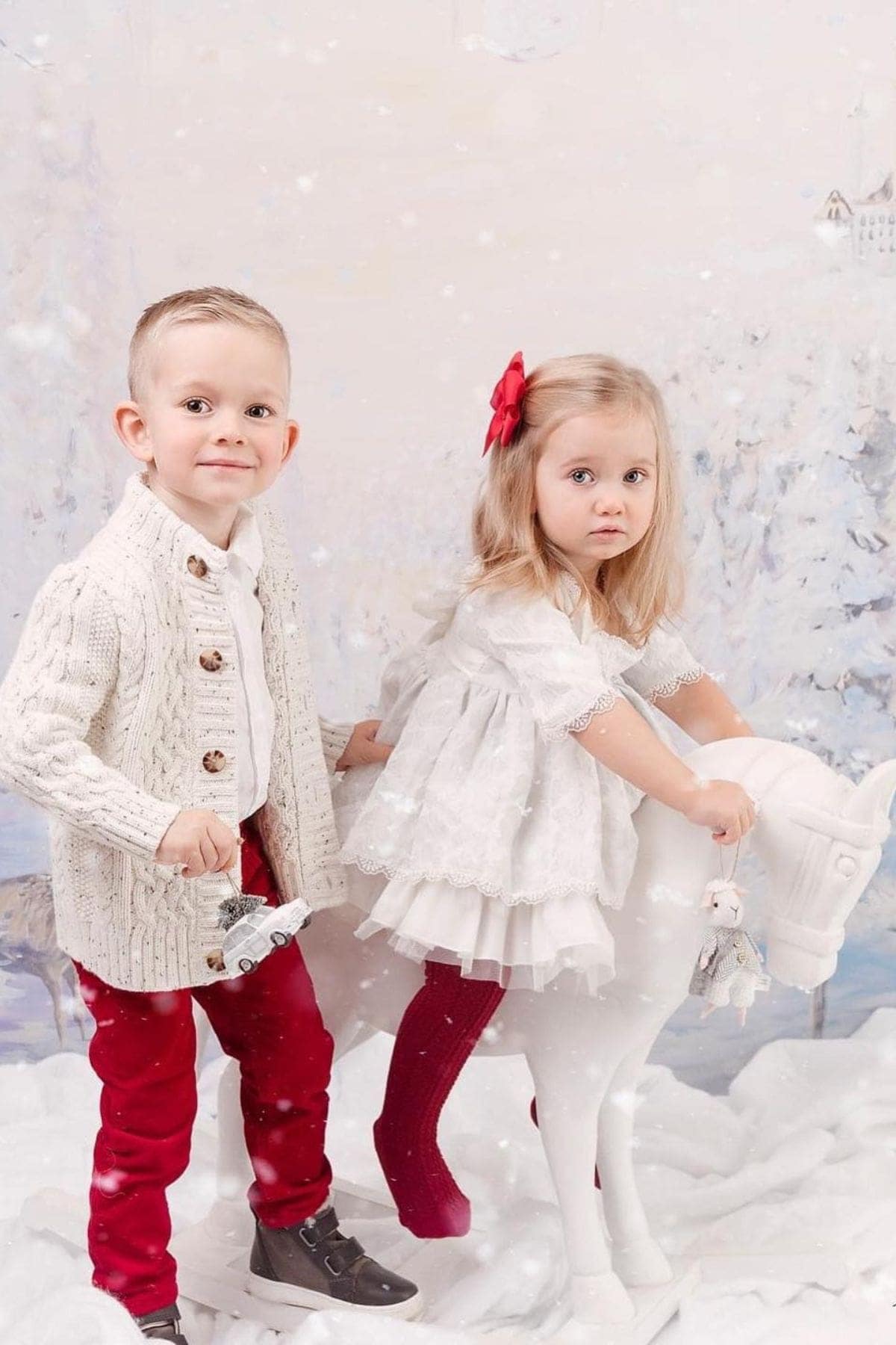 A young boy in red pants and a girl in a white dress with a red bow stand by a white toy horse in a snowy, winter-themed setting. Snowflakes appear to fall around them.