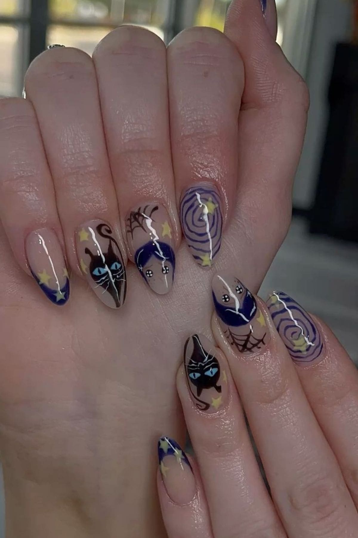 Hands with long nails feature a Halloween-themed manicure, including black cat faces, stars, spider webs, and spiral designs in dark blue, beige, and yellow colors.