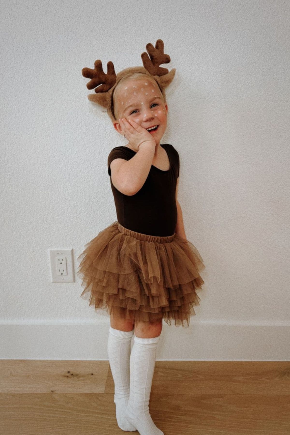 Young child dressed in a brown tutu, black leotard, white knee-high socks, and antler headband poses and smiles against a plain wall.