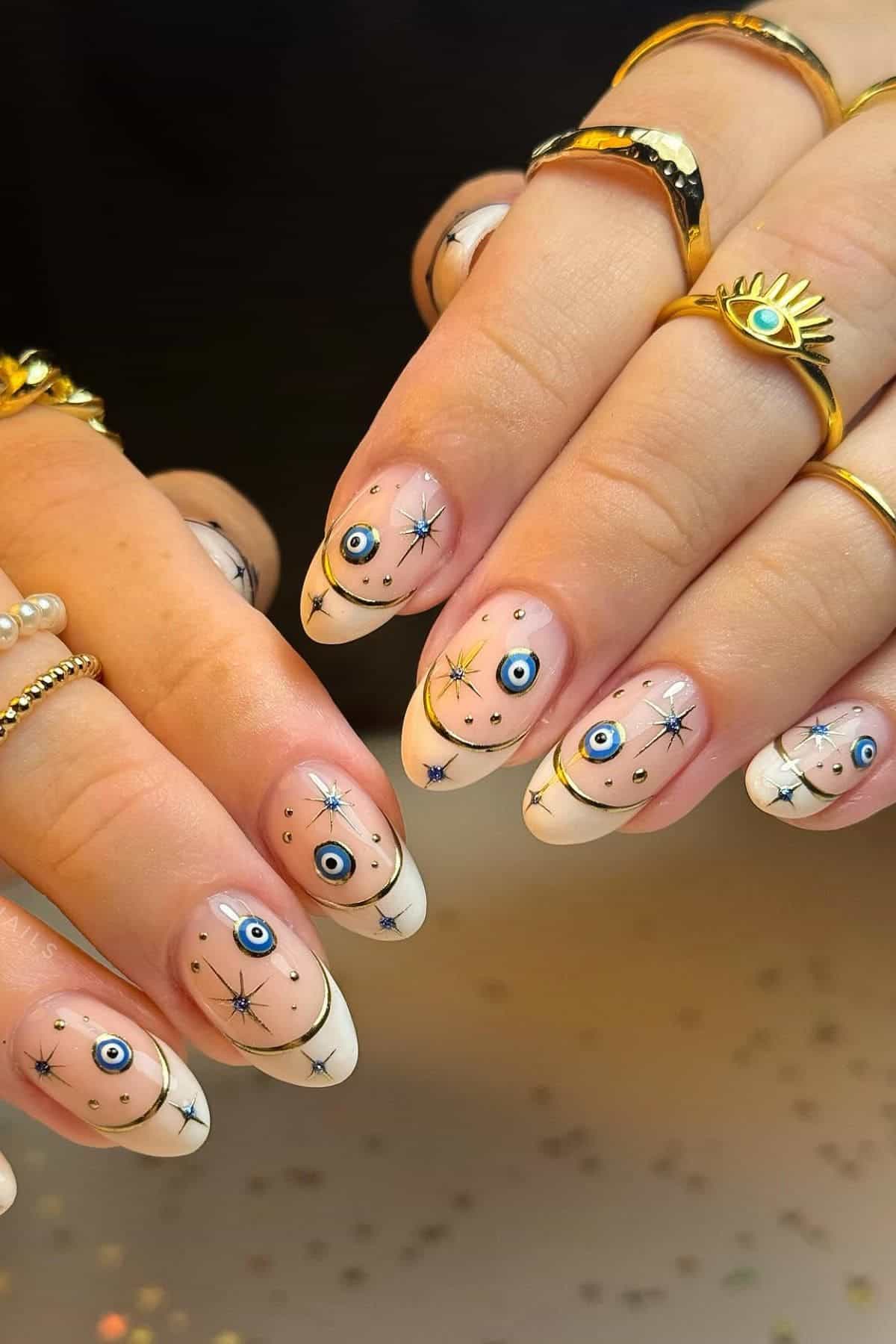 Hands with almond-shaped nails featuring white French tips and blue evil eye and star designs; gold rings and bracelets are worn on fingers.