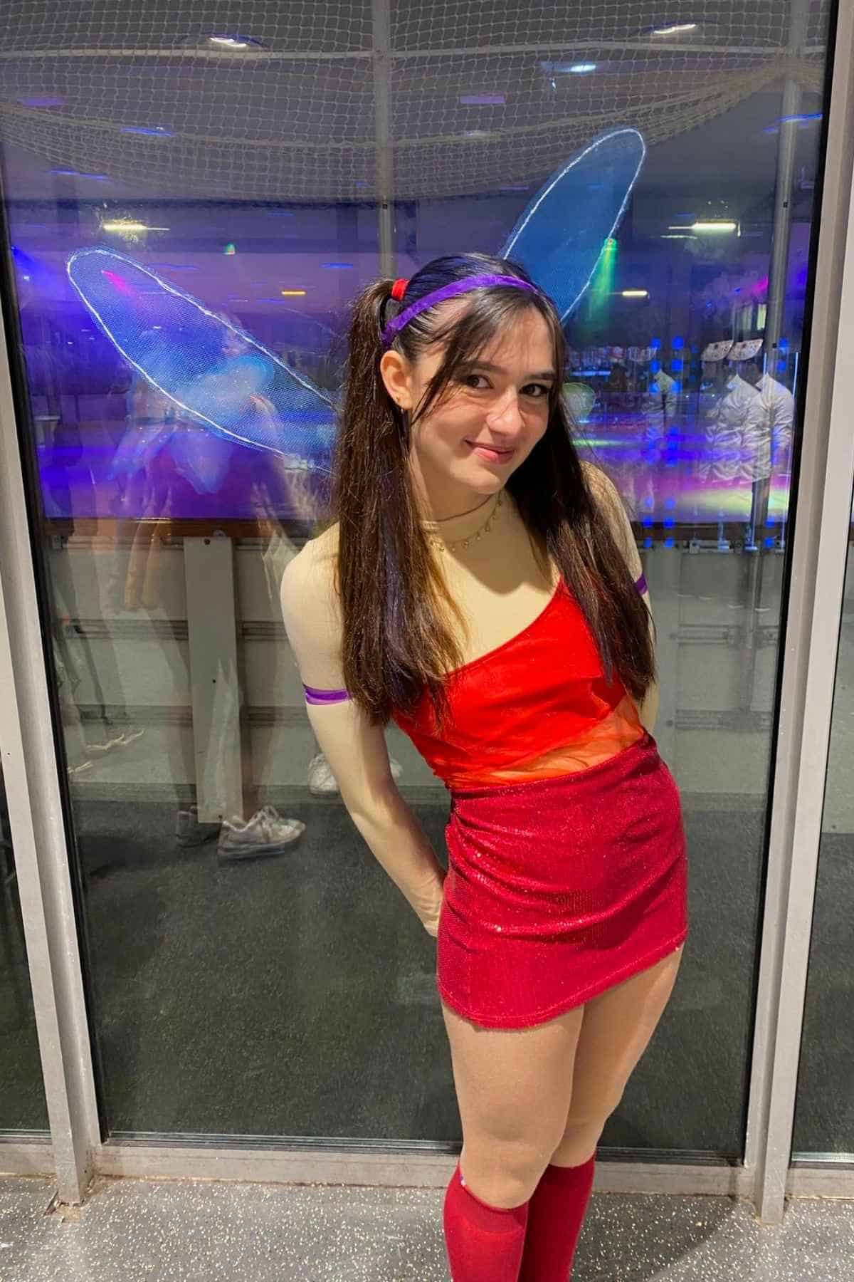 Person wearing blue fairy wings, a red one-shoulder dress, and red knee-high socks stands indoors in front of glass doors, smiling at the camera.