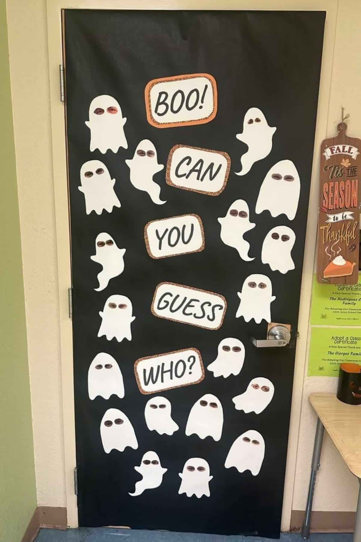 A classroom door decorated with white cartoon ghosts and signs that read, “Boo! Can you guess who?” on a black background.