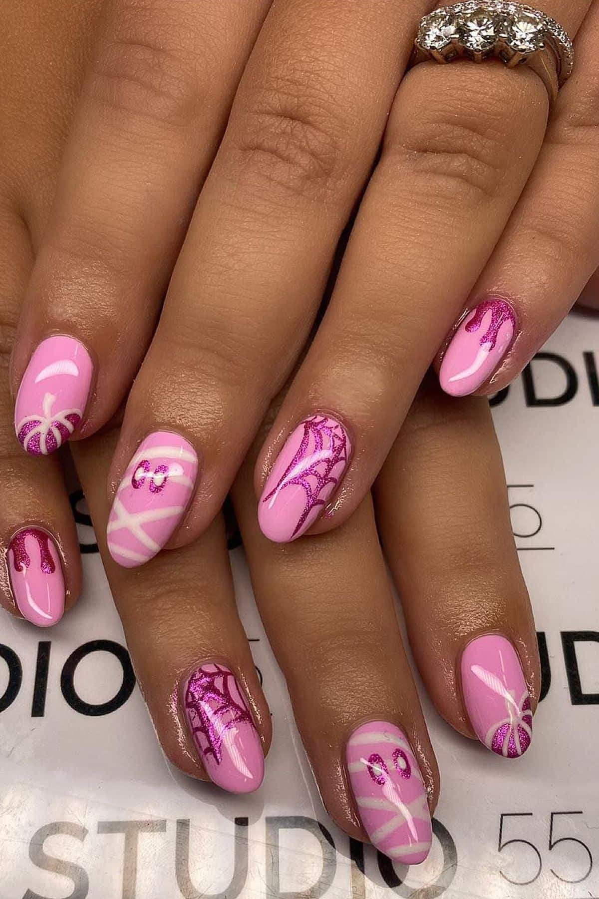 Hands with pink Halloween-themed nail art featuring spider webs, a spider, and dripping designs; a diamond ring is on one finger.