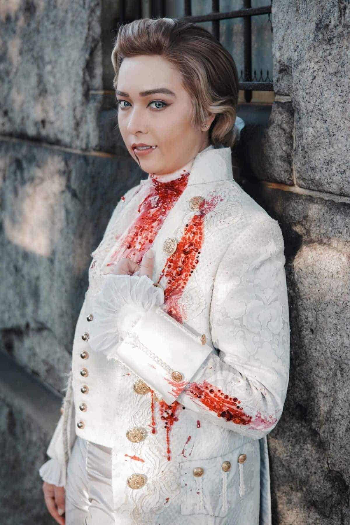 Person in an ornate white historical costume with gold buttons, posing against a stone wall, with red fake blood on their neck and chest.