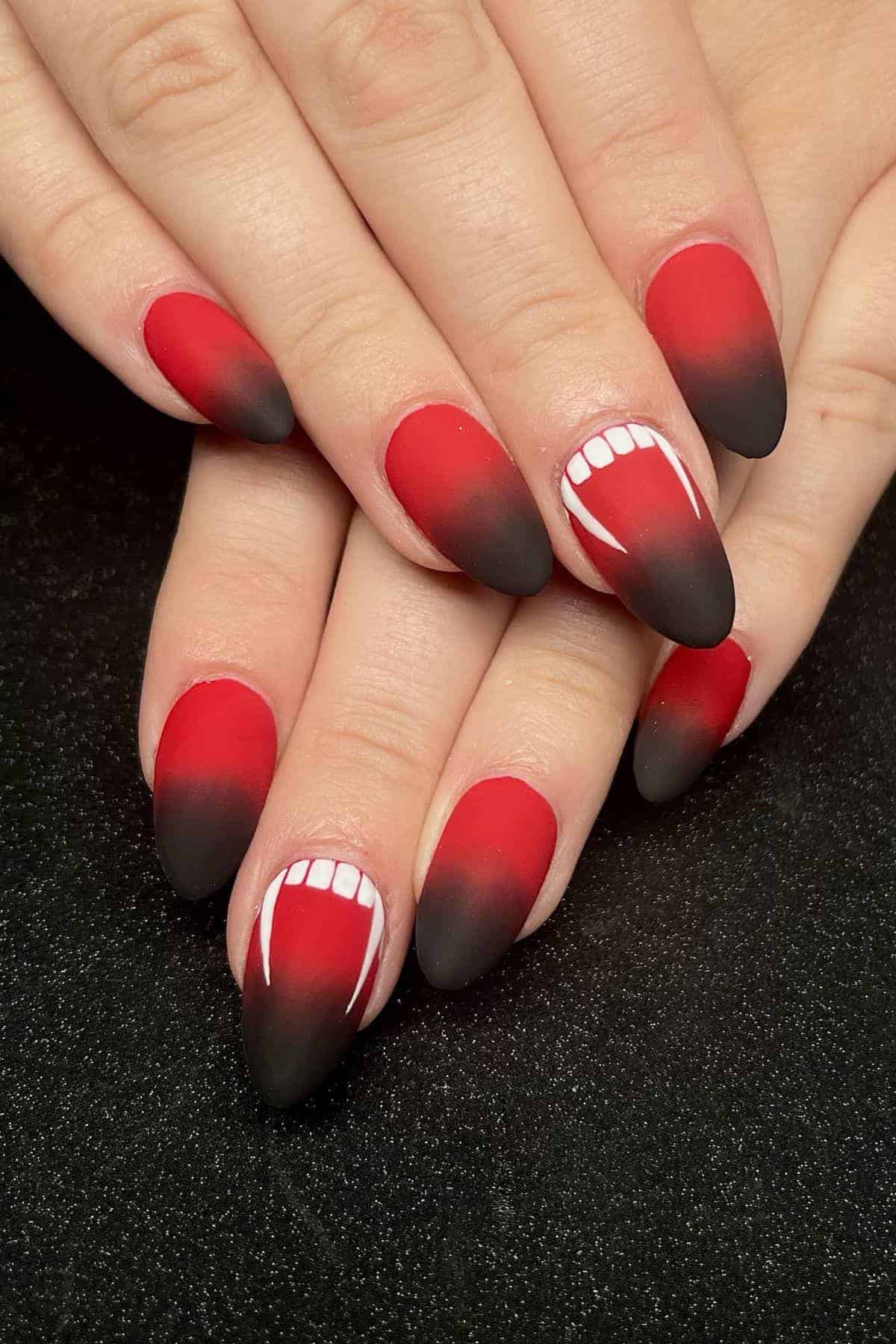 Hands with red and black ombre almond-shaped nails, featuring white fang and teeth designs on two nails, displayed against a black background.