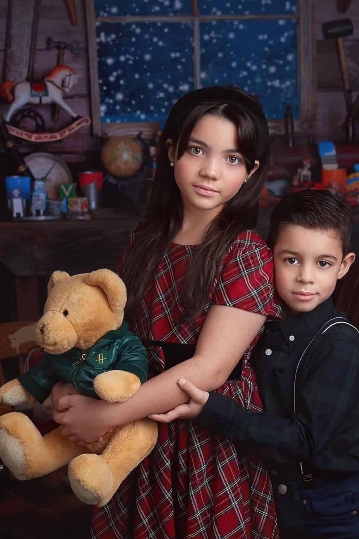 Two children pose indoors; the older girl holds a teddy bear, and a younger boy stands beside her. A snowy window, toys, and books are visible in the background.