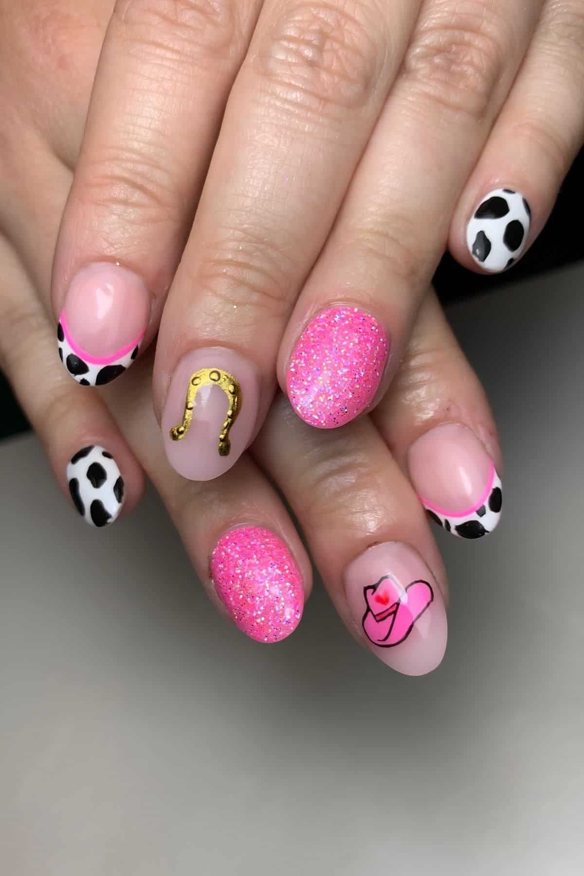 Hands with manicure featuring pink glitter, cow print, gold horseshoe, and a pink cowboy hat design on oval-shaped nails.