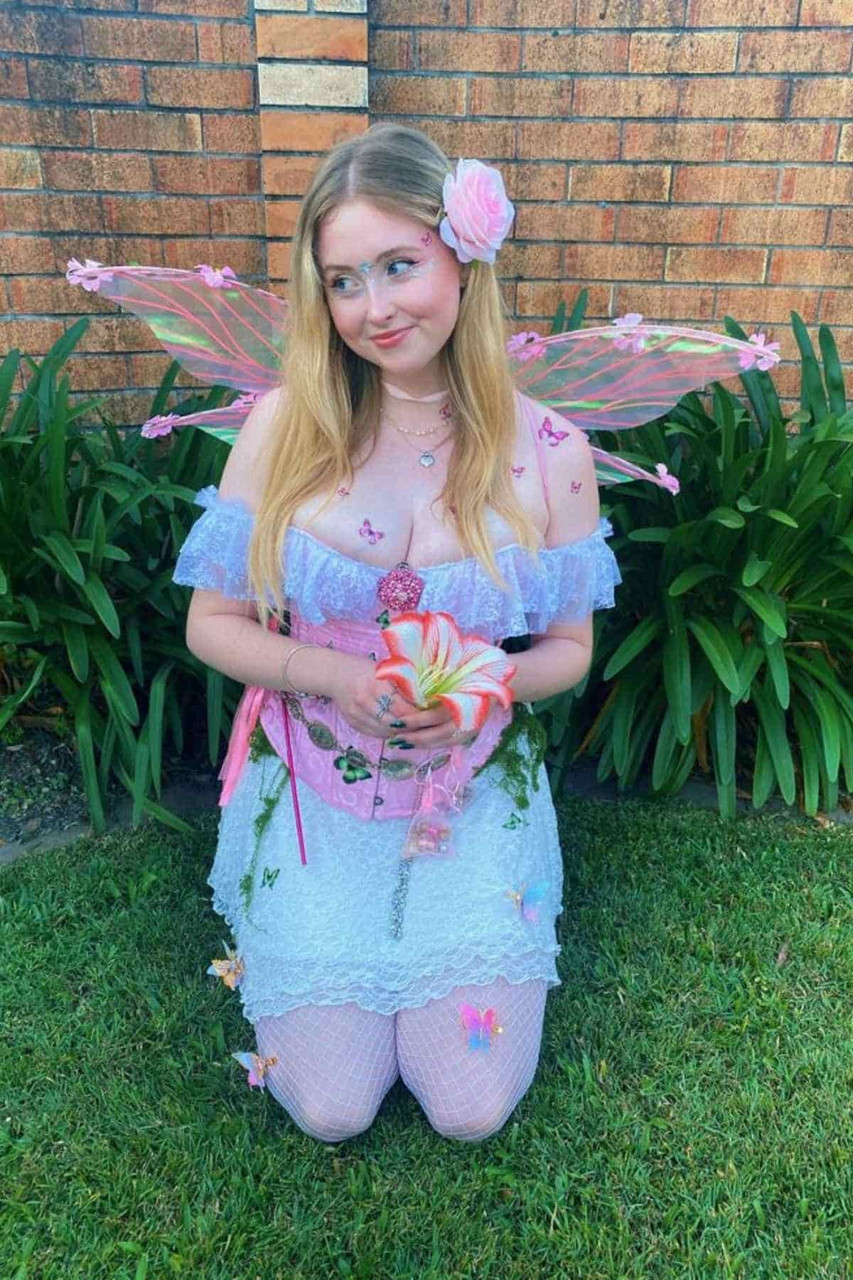 Person in a fairy costume with pink wings, a flower in their hair, and holding a flower, kneels on grass in front of a brick wall with green plants.