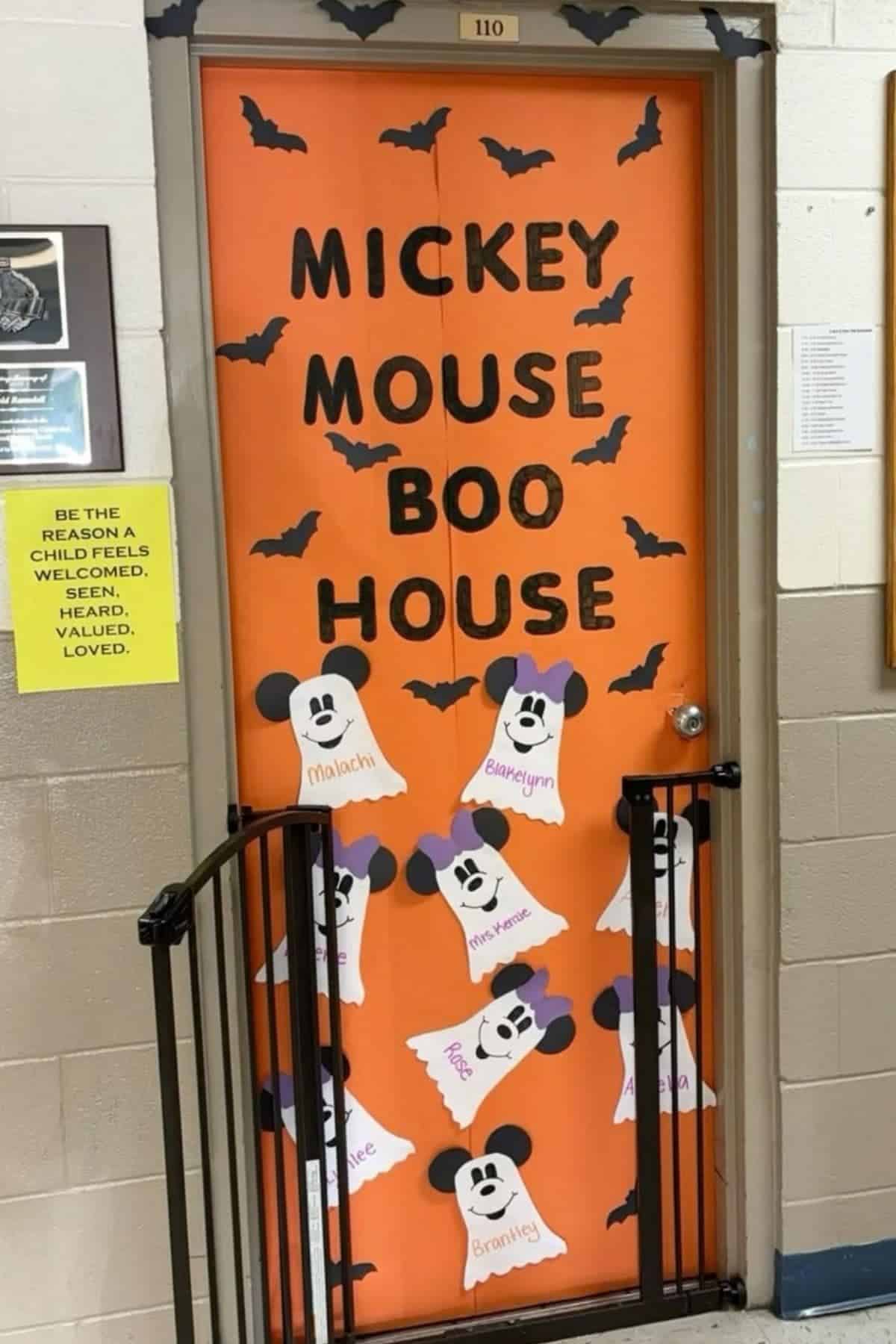 An orange classroom door reads “Mickey Mouse Boo House,” decorated with black bats and paper ghosts featuring Mickey Mouse faces and names written on them.