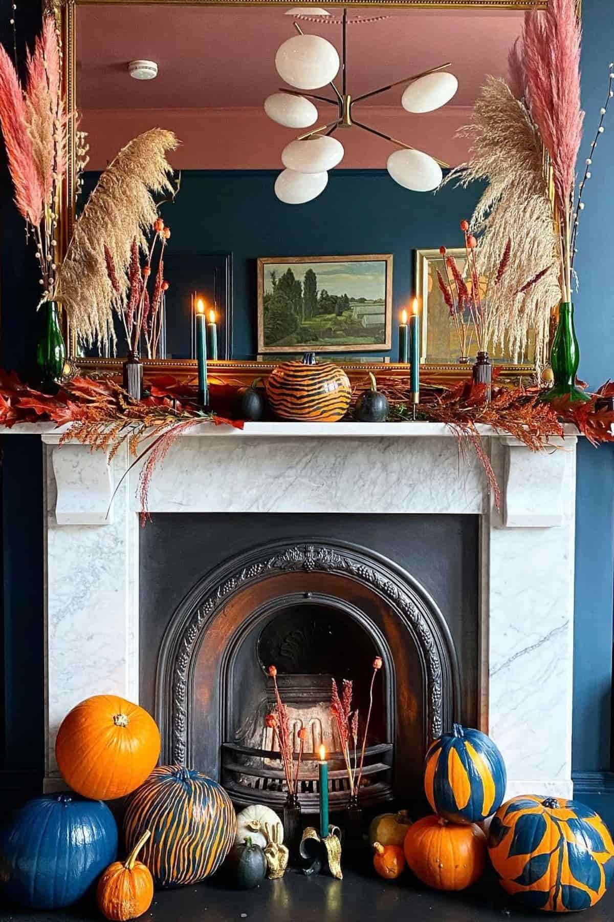 A marble fireplace decorated with painted pumpkins, candles, pampas grass, and autumn leaves, with framed art and a mirror above the mantel.