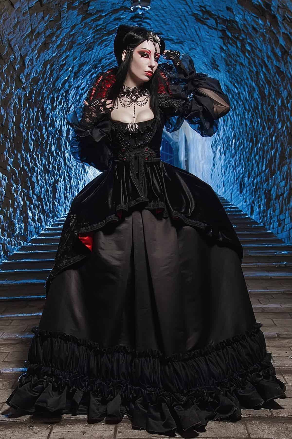 Person in a long black gothic dress with sheer sleeves and dramatic makeup stands in a stone tunnel with blue lighting.