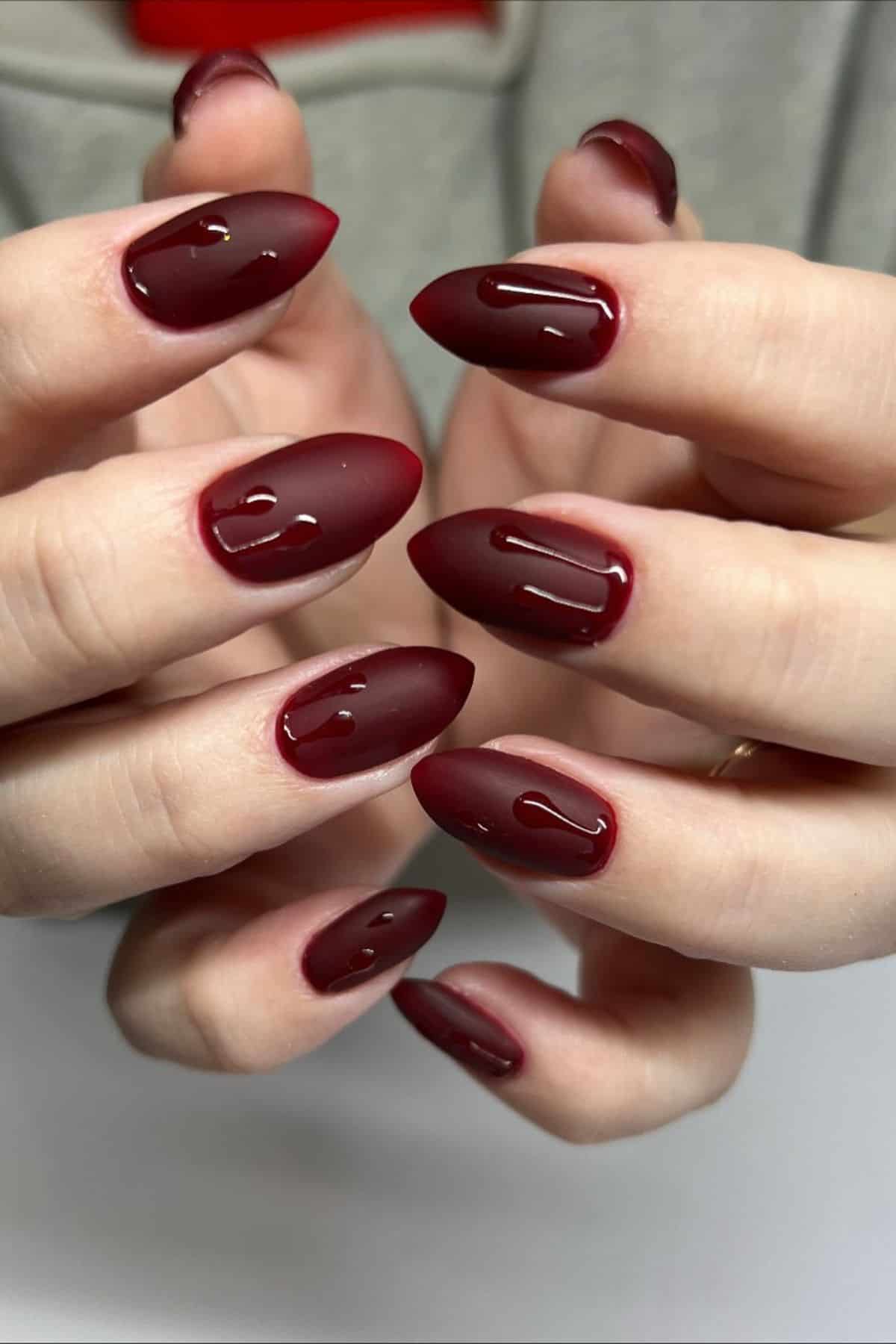 Close-up of hands with almond-shaped nails painted matte burgundy and glossy drips on each nail, resembling a liquid effect.