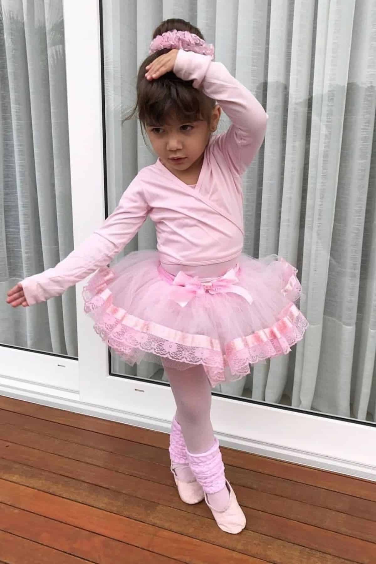 Young child in a pink ballet outfit and tutu poses with one arm raised, standing on a wooden floor in front of glass doors with white curtains.