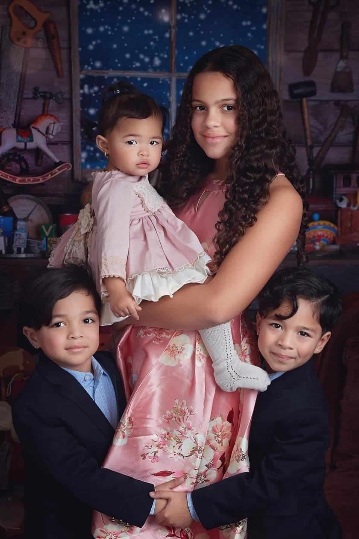 Four children in formal attire pose together indoors with a rustic, toy-filled background and a window showing a starry night sky.