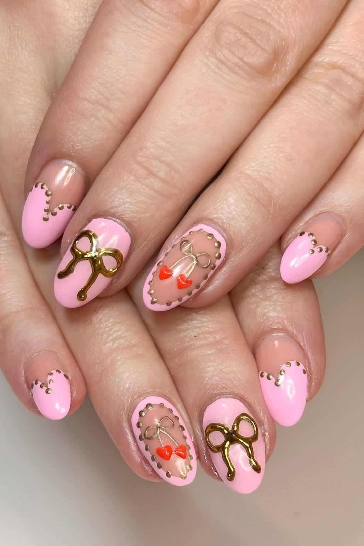 Hands with almond-shaped nails painted pink, decorated with gold bows, heart designs, and gold dots along the cuticle line.
