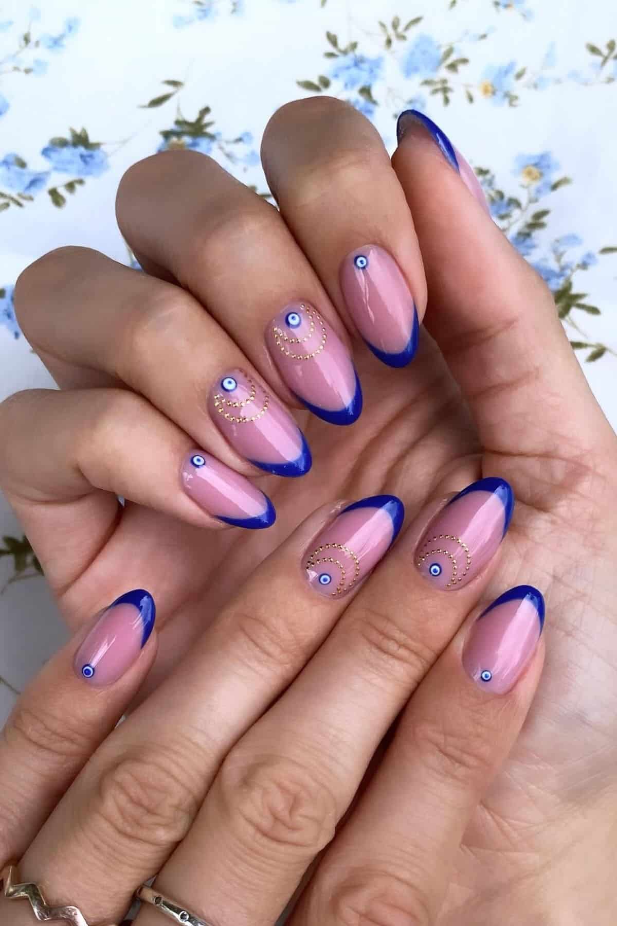 Hands with almond-shaped nails, pink base, blue tips, and gold designs with small blue and white circles. Background is a floral fabric.