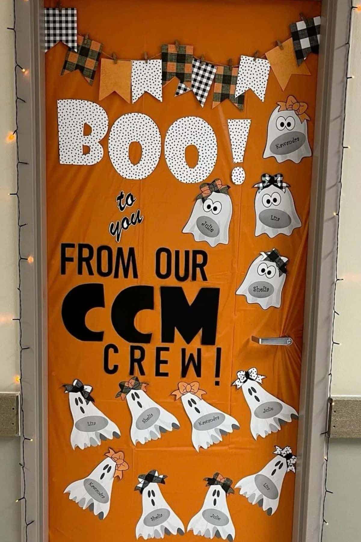 An orange door decorated with “BOO! From our CCM crew!” and cutout ghosts labeled with names, festive banners, and string lights.