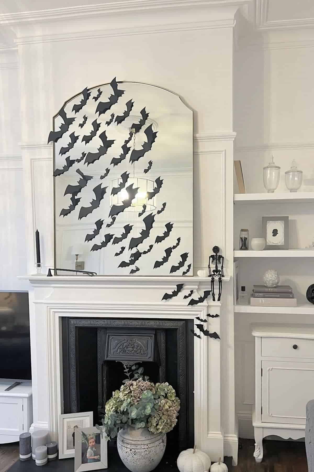 A large mirror above a white fireplace is decorated with black paper bats. The mantel holds candles, a nutcracker figure, and dried flowers. White shelves and décor fill the background.