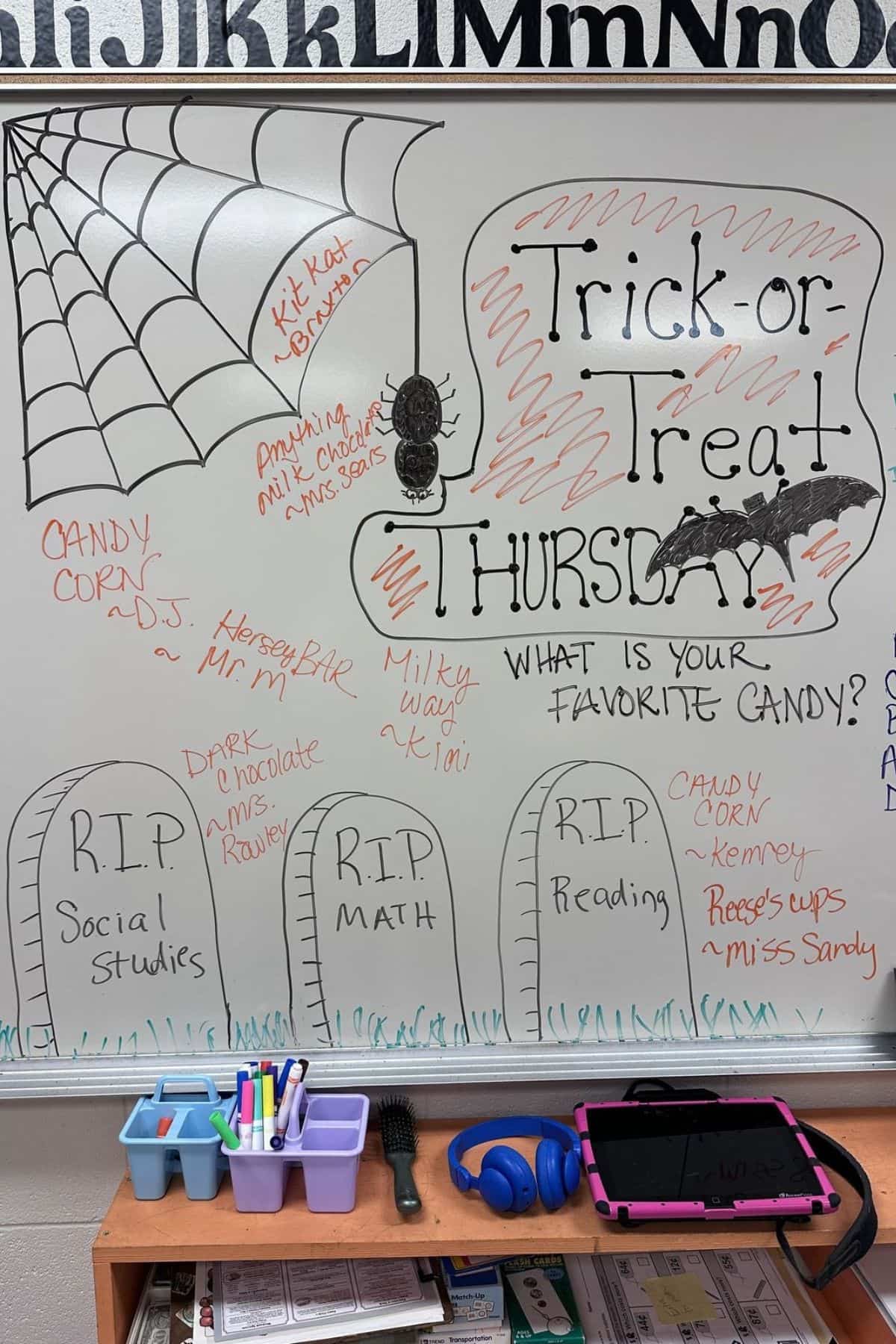 Whiteboard with Halloween drawings, candy names, and “Trick-or-Treat Thursday.” Sections labeled RIP for Social Studies, Math, and Reading. Markers, supplies, and a tablet are on a tray below.