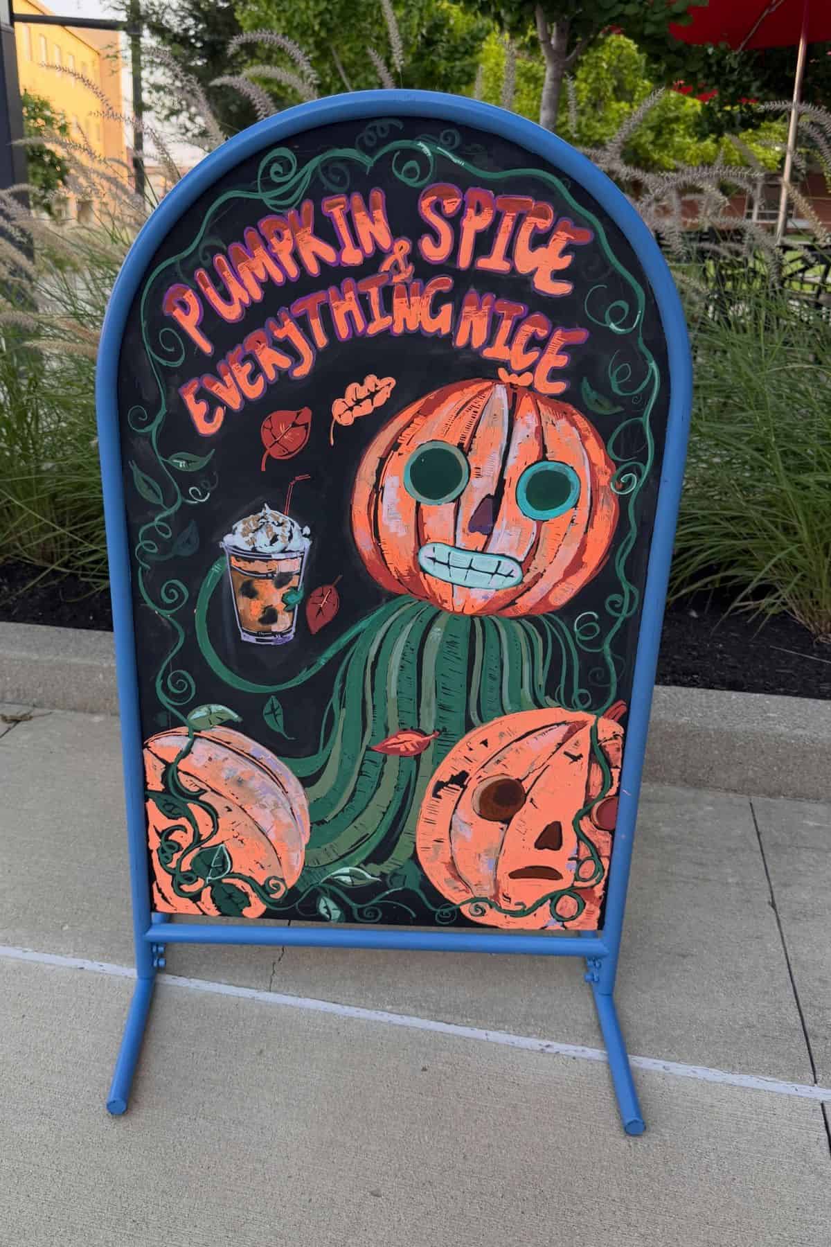 Chalkboard sign with pumpkin drawings and a pumpkin spice drink, reading “Pumpkin Spice & Everything Nice,” placed on a sidewalk near grass and plants.