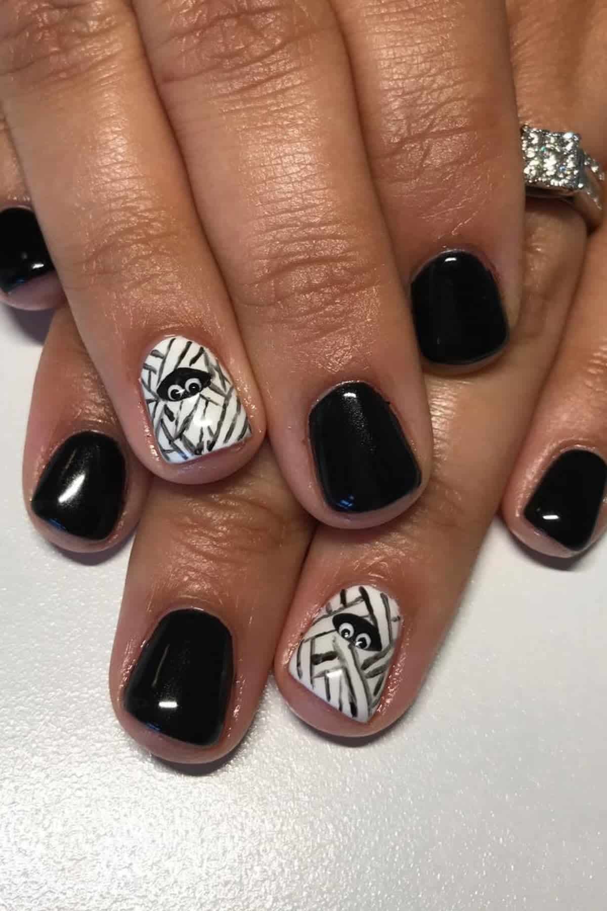 Hands with black nail polish; ring fingers have white and black mummy designs with cartoon eyes.