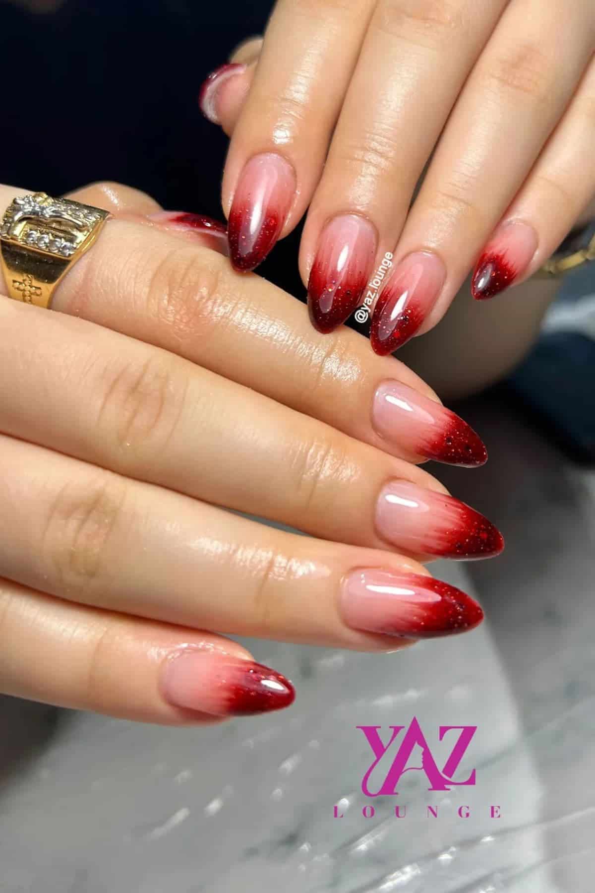Hands with almond-shaped nails featuring a red ombre design, fading from dark red tips to a natural base. A gold ring is worn on one finger. “YAZ LOUNGE” logo visible at the bottom.