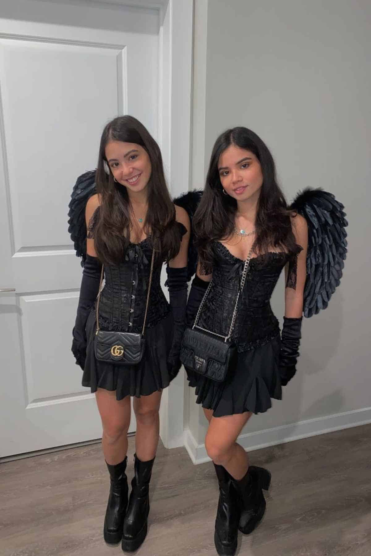Two young women pose indoors wearing matching black outfits with corsets, skirts, gloves, boots, black angel wings, and crossbody bags, standing in front of a white door and wall.