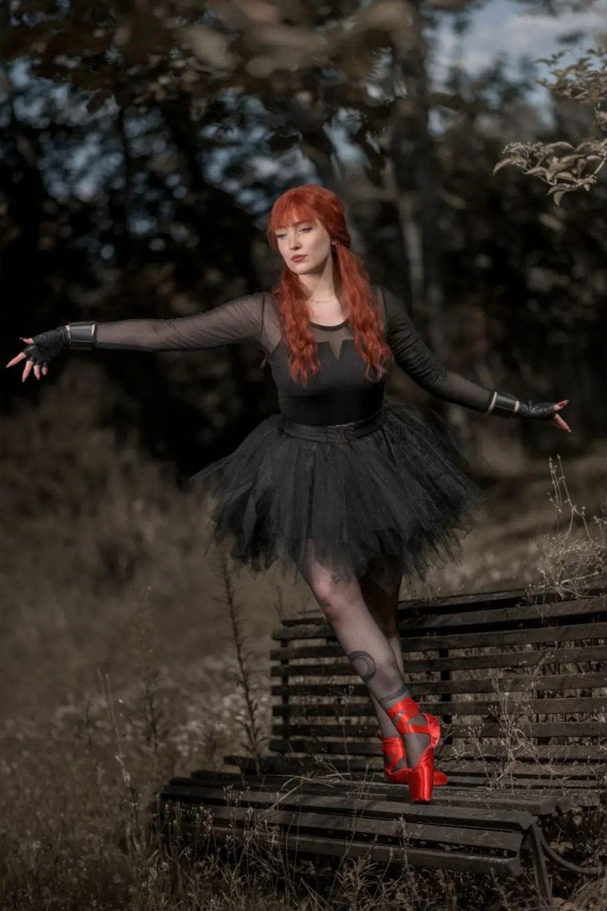 A woman with long red hair in a black tutu and red pointe shoes poses on a wooden bench outdoors, surrounded by foliage.