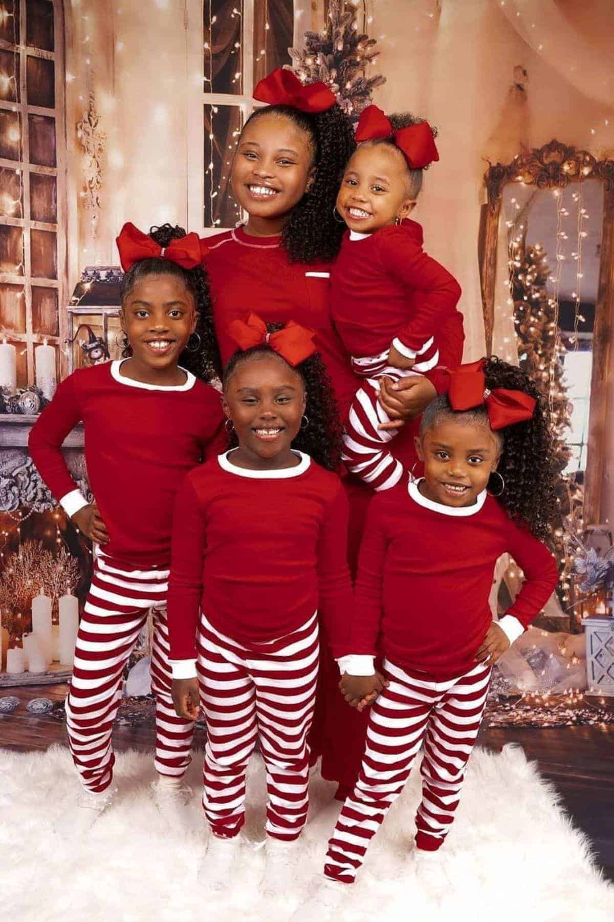 Five girls pose in matching red tops and red-and-white striped pants with big red bows in their hair, standing on white fur in a festive holiday setting with Christmas lights and decorations.