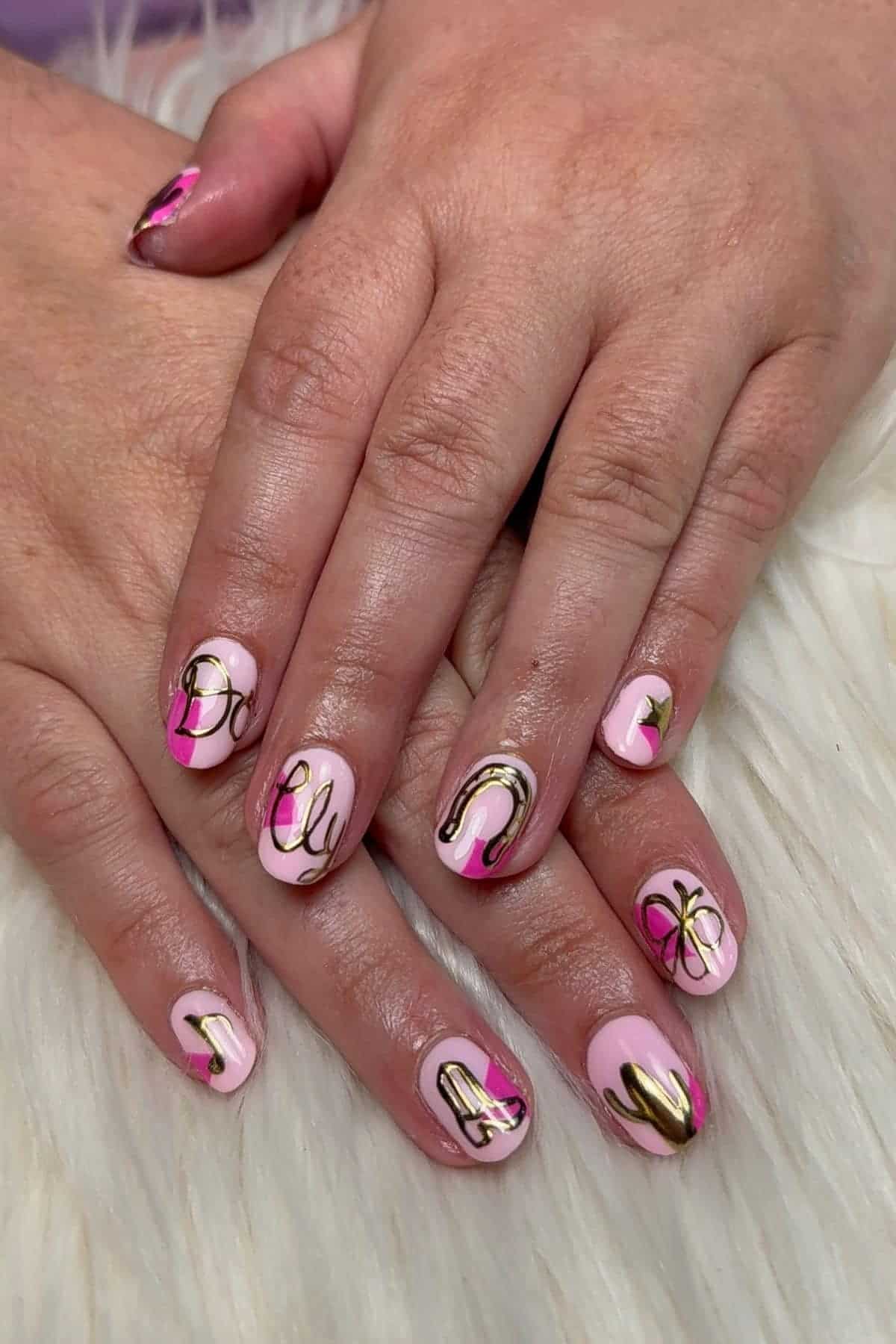 Hands with pink and gold nail art. Each nail has unique abstract designs and gold outlines, with some nails featuring stylized letters. The hands rest on a white, fluffy surface.