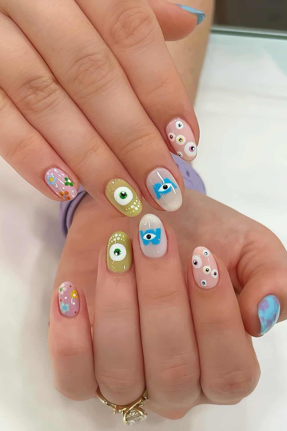 Hands with pastel-colored nail art featuring various eye designs, polka dots, and a blue sky motif on the pinky finger.