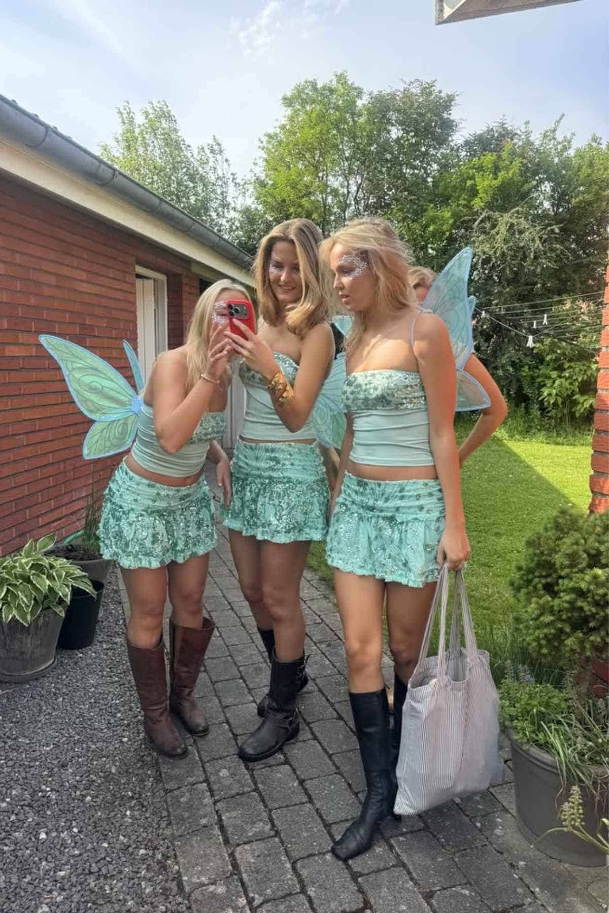 Four people dressed in matching mint green fairy costumes stand outside on a stone path; one person takes a selfie while the others pose.