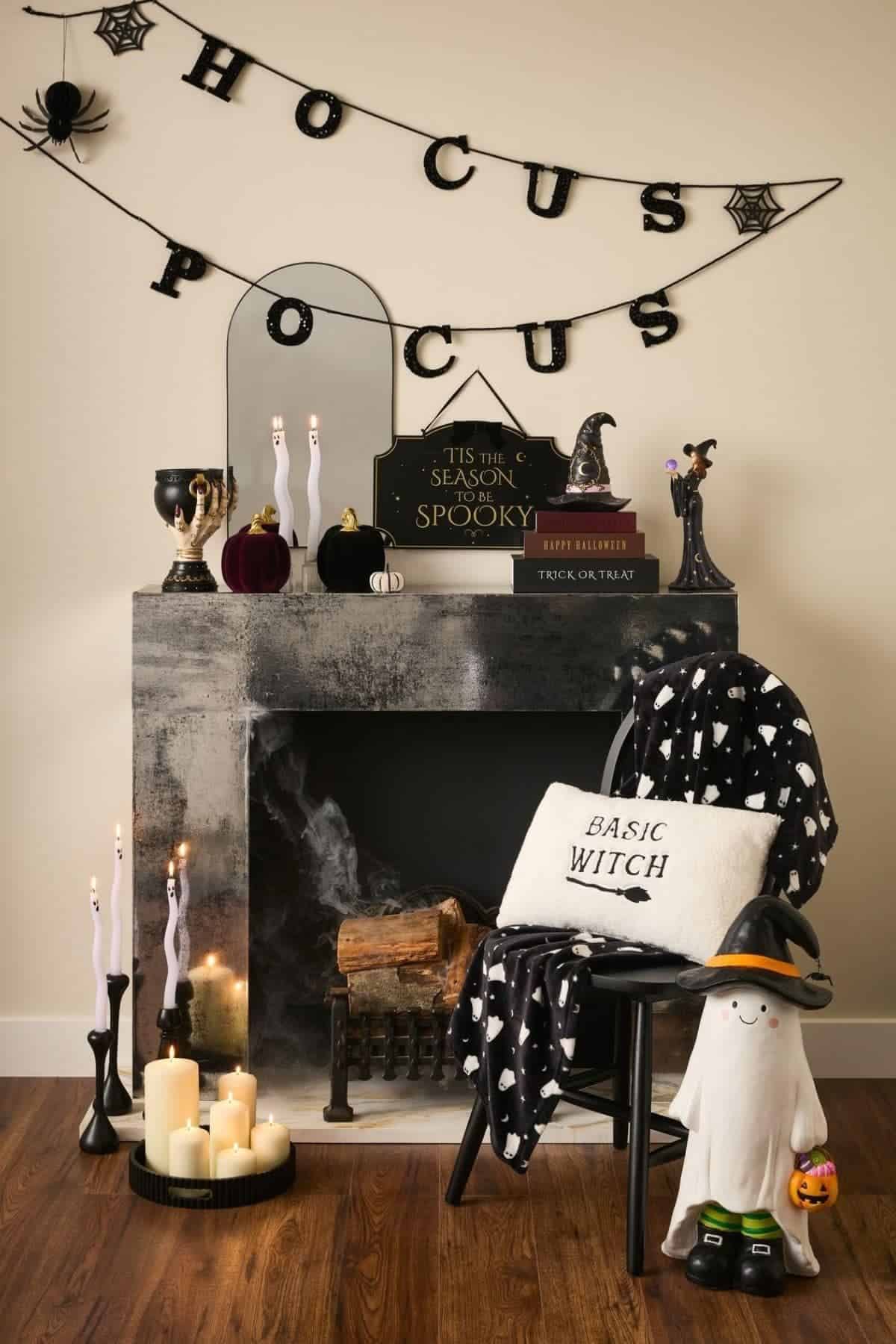 A Halloween-themed room with a faux fireplace, candles, black cat and witch decor, a “HOCUS POCUS” banner, and a ghost figure holding a pumpkin.