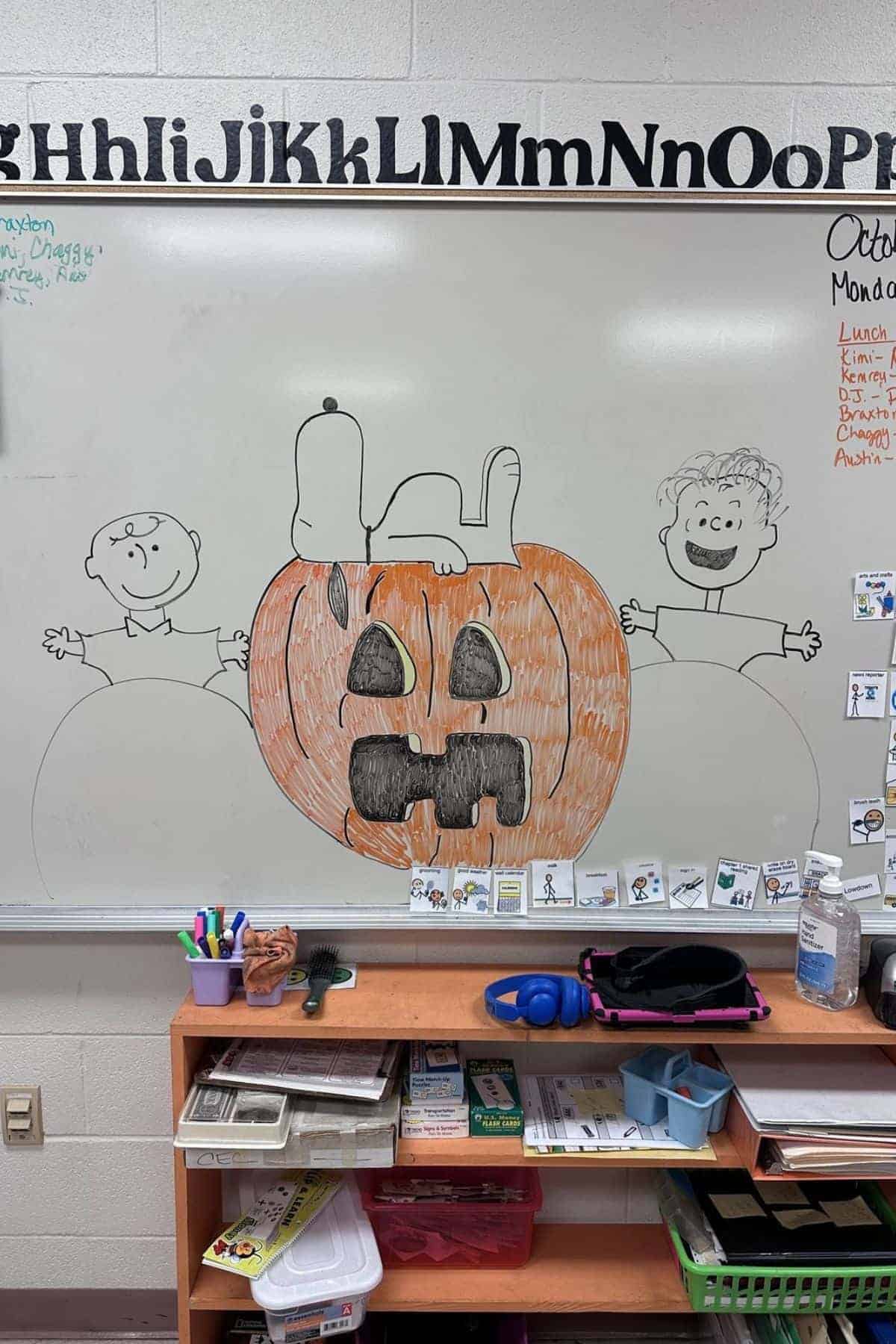 Whiteboard drawing of a pumpkin with a Jack-o’-lantern face, two cartoon children, and a dog on top. Alphabet letters border the top. Classroom supplies and a shelf are below the board.
