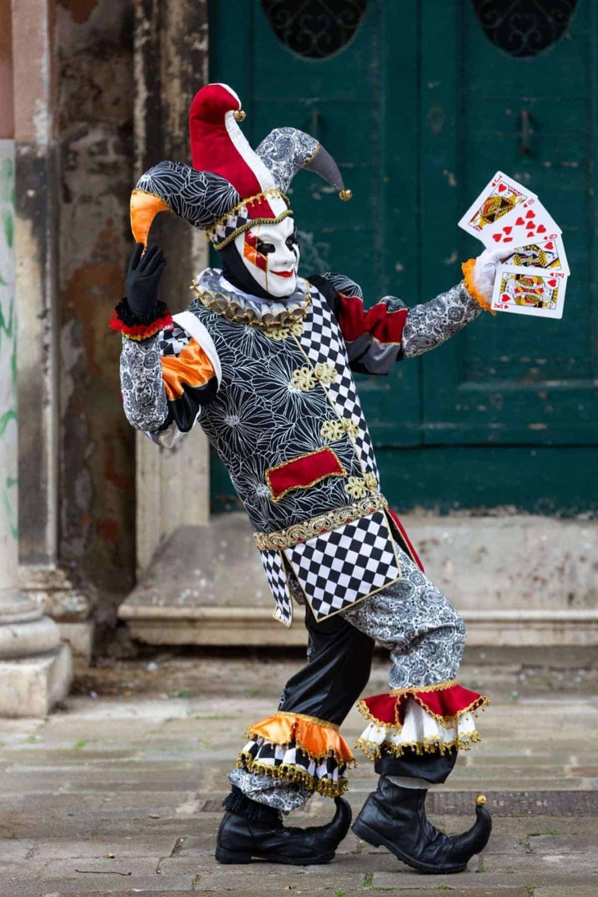 A person in an elaborate jester costume poses outdoors, holding playing cards in one hand.